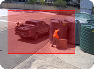 Person in an orange vest unloading a box from a pickup truck near large green storage tanks in a paved outdoor area.