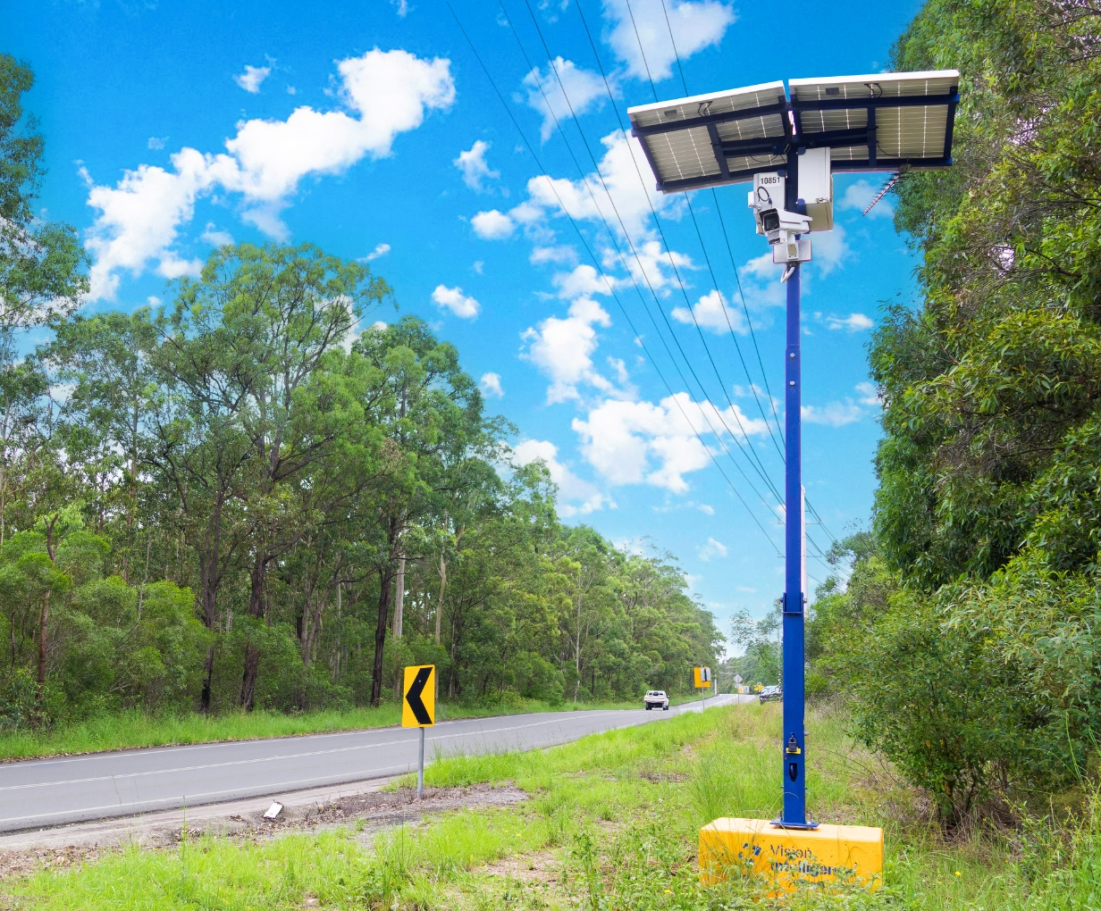 Solar-powered speed camera mounted on a blue pole beside a rural road with green trees and cloudy blue sky.