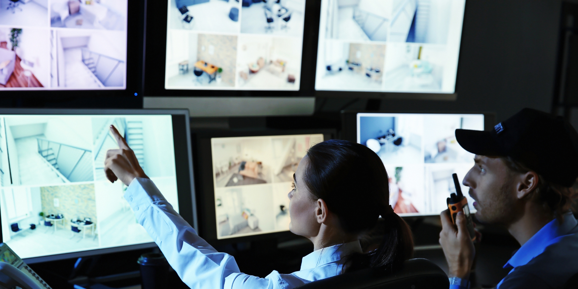 Two security personnel monitoring multiple CCTV screens showing indoor surveillance footage.