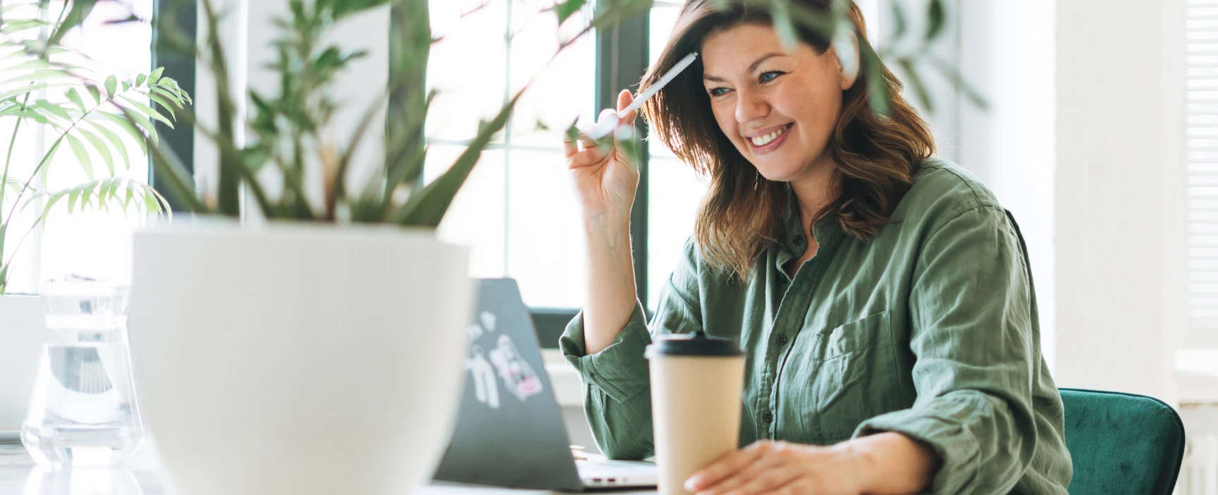 Smiling woman with brown hair, wearing a green shirt, sitting at a table with a laptop and a coffee cup, holding a pen near her temple, in a bright room with plants and large windows.