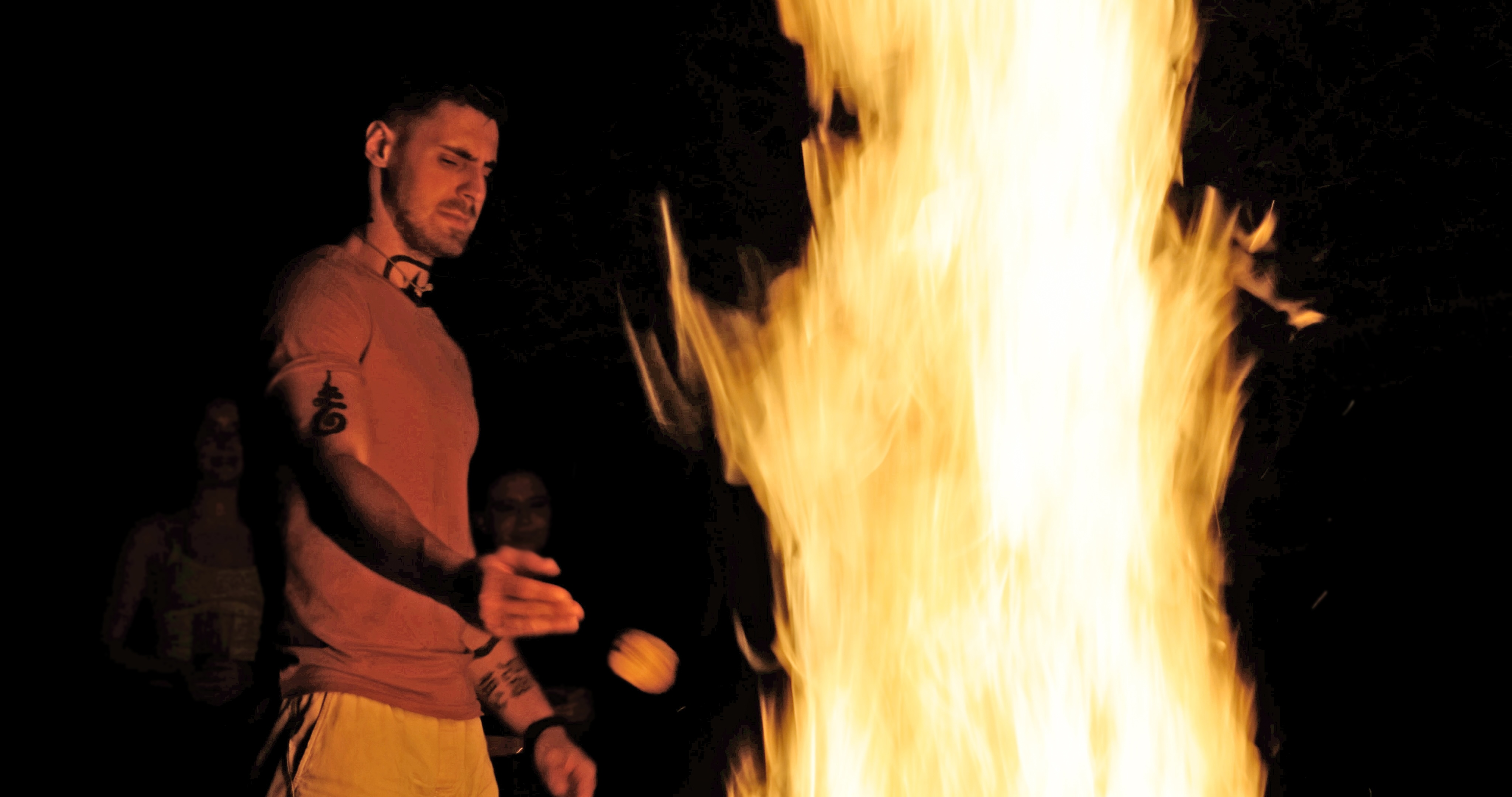 Man with tattoos roasting marshmallow over a large campfire at night with two people faintly visible in the background.