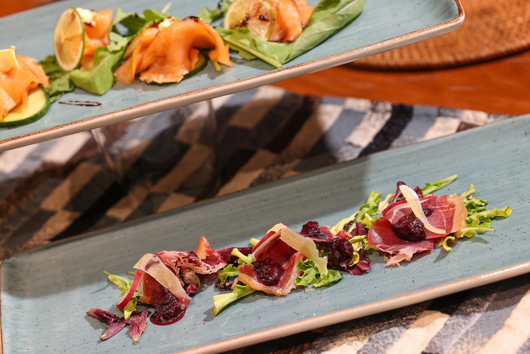 Blue rectangular plate with three servings of thinly sliced cured meat garnished with greens, shaved cheese, and dark dried fruits.
