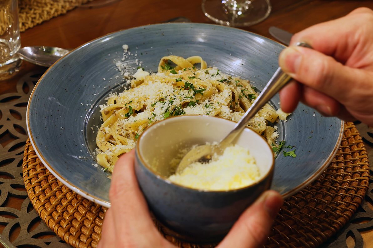 Hand sprinkling grated cheese from a small bowl onto a plate of pasta garnished with herbs.