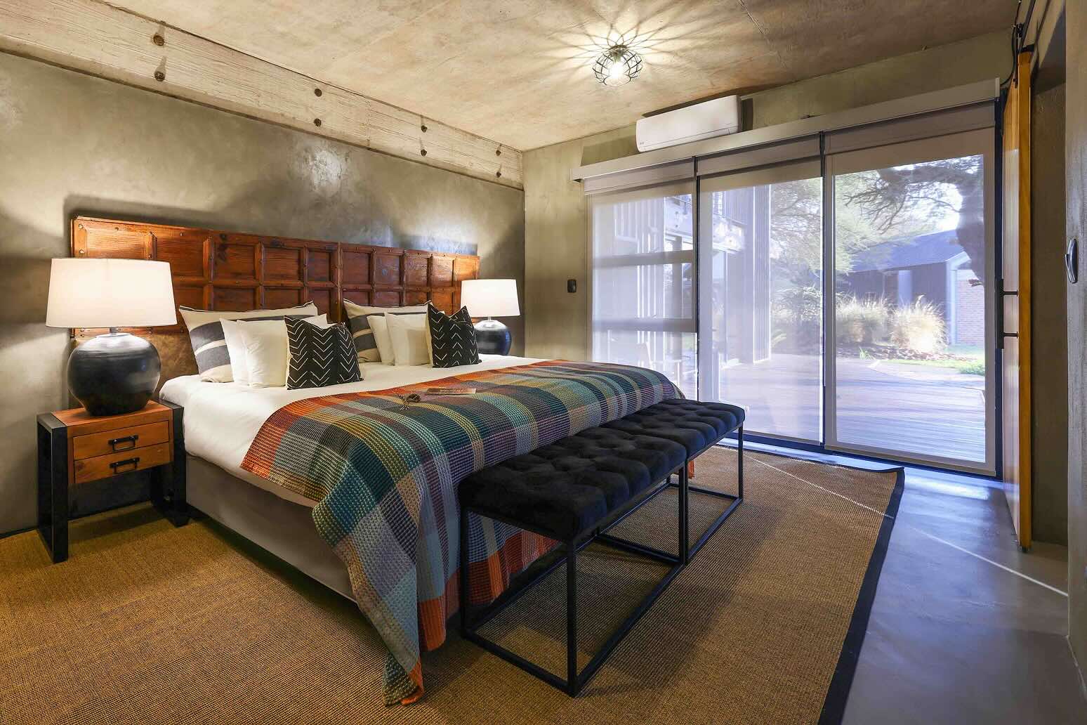 Naka Celestial Suites. Modern bedroom with a large bed featuring a wooden headboard, patterned throw, two bedside lamps, and sliding glass doors with a view outside.