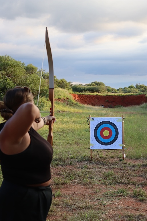 Person aiming a bow and arrow at a colorful archery target set in a grassy outdoor range.