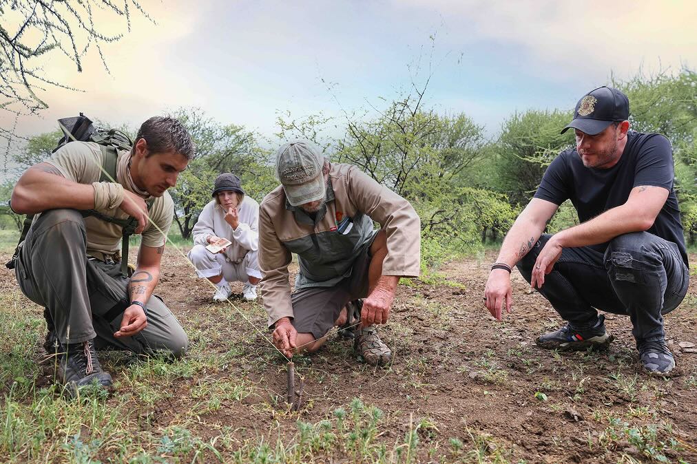 Four people crouched outdoors on dirt and grass, examining a small wooden stake tied with string in the ground amid sparse bushes.