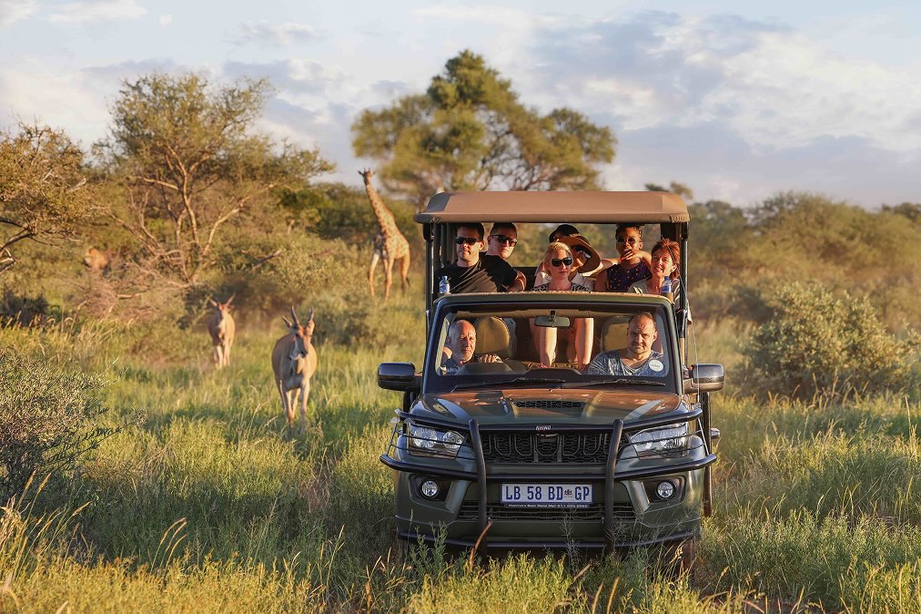 Group of people on a safari vehicle observing wildlife including giraffe and antelopes in a grassy savanna.