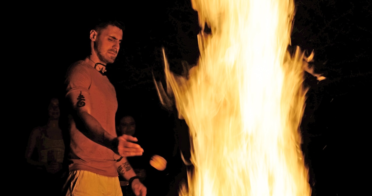Man with tattoos roasting marshmallow over a large campfire at night with two people faintly visible in the background.