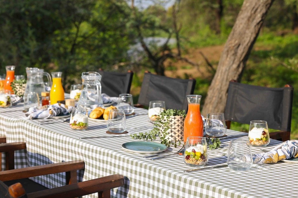 Outdoor dining table with green and white checkered tablecloth, glasses of fruit parfait, pitchers of juice and water, croissants, and black chairs in a garden setting.