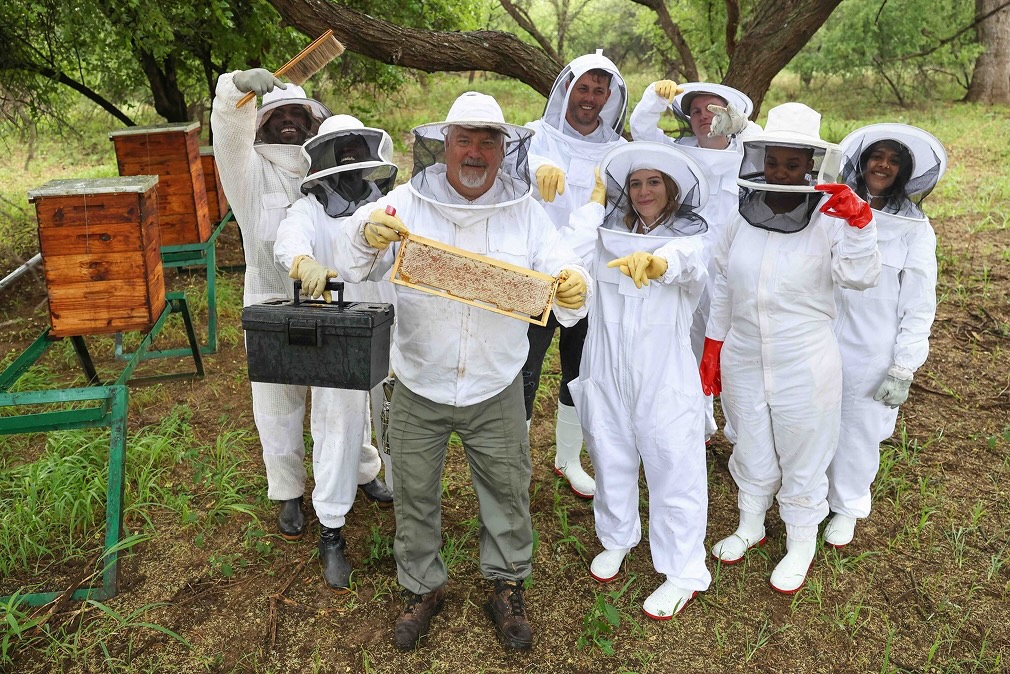 Group of beekeepers in protective suits standing outdoors next to wooden beehives, holding honeycomb frame and beekeeping tools at Naka Luxury Lodge for Hunting for Honey Event.