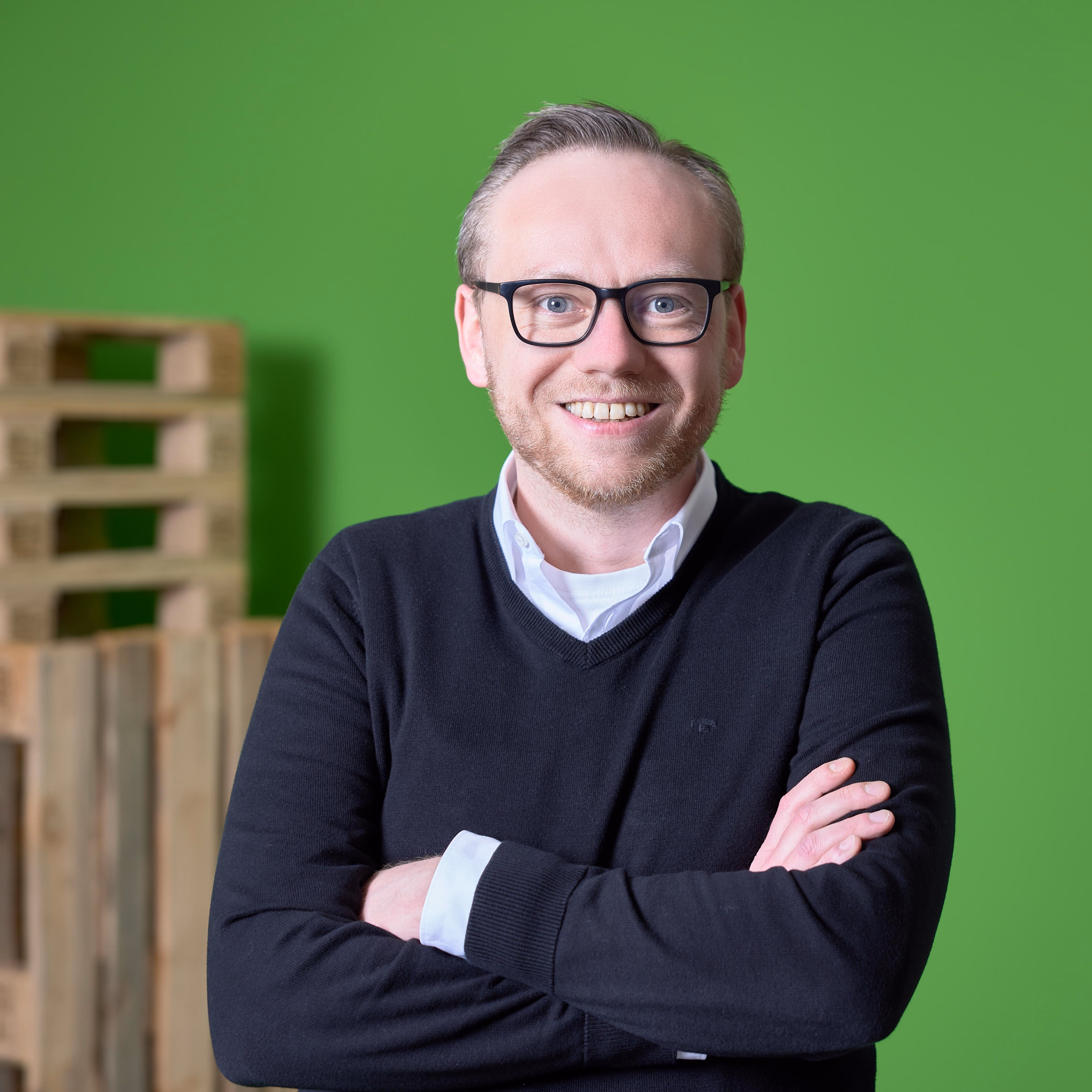Lächelnder Mann mit Brille und schwarzen Pullover vor grünem Hintergrund mit Holzpaletten.