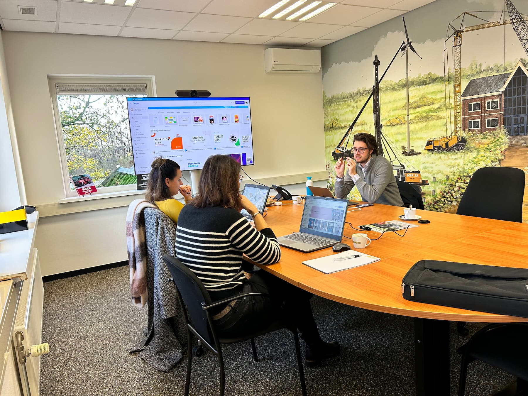 Three people in a conference room with laptops discussing in front of a screen and a mural of cranes.