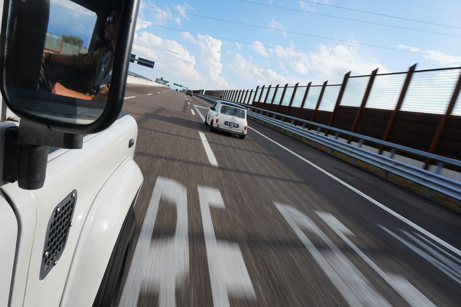 Rear view of a white vintage car driving on the highway in front of the vehicle with side mirror reflection.