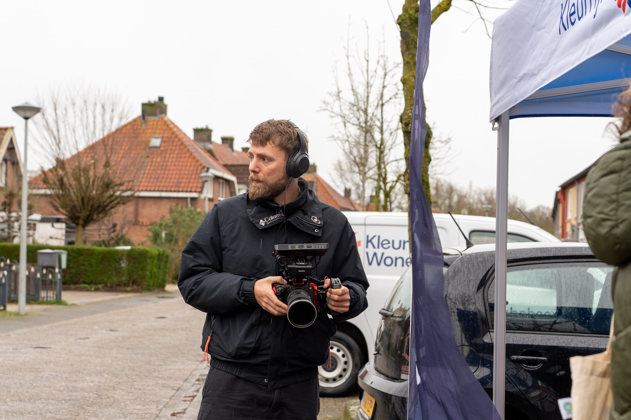 Man wearing headphones and black jacket operates a video camera outdoors on a cloudy day.