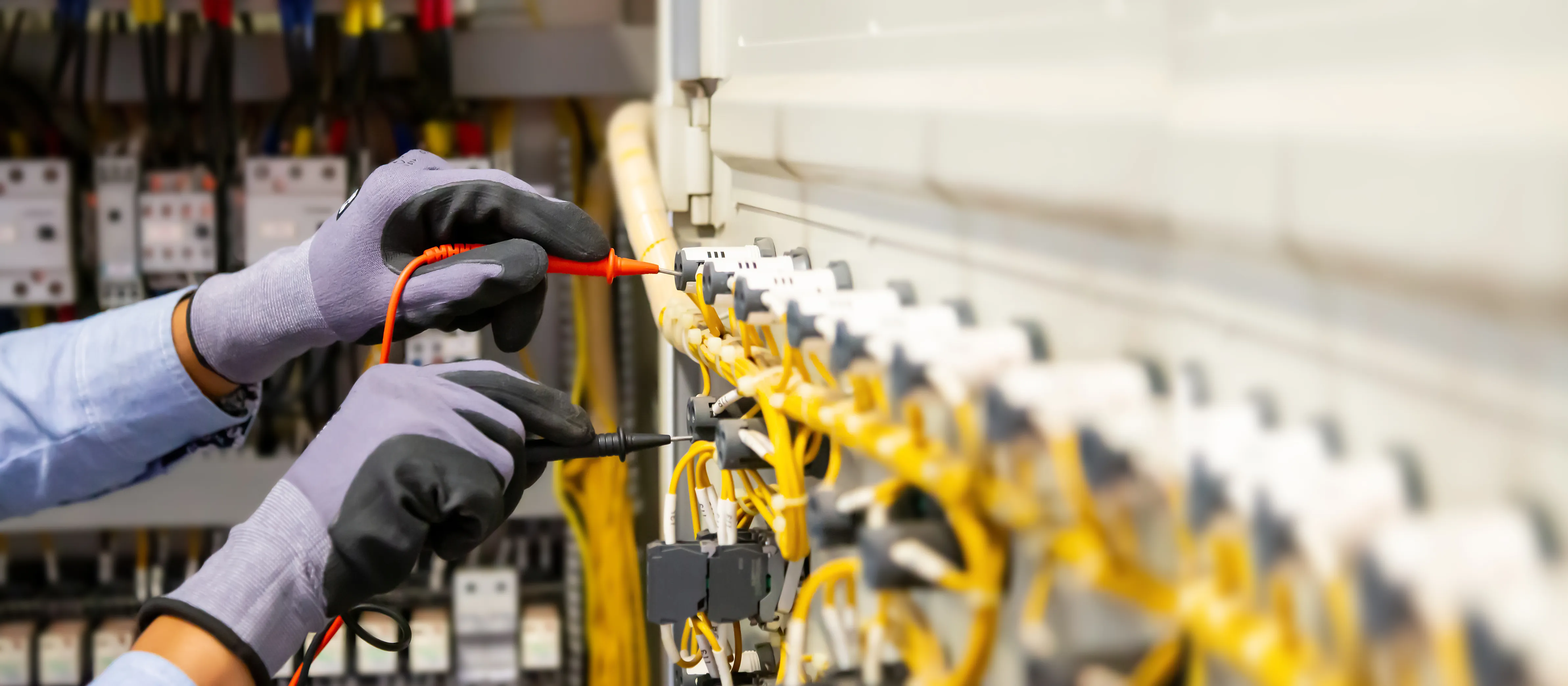 Person wearing gloves and using a multimeter to test electrical connections in a control panel with numerous yellow wires.