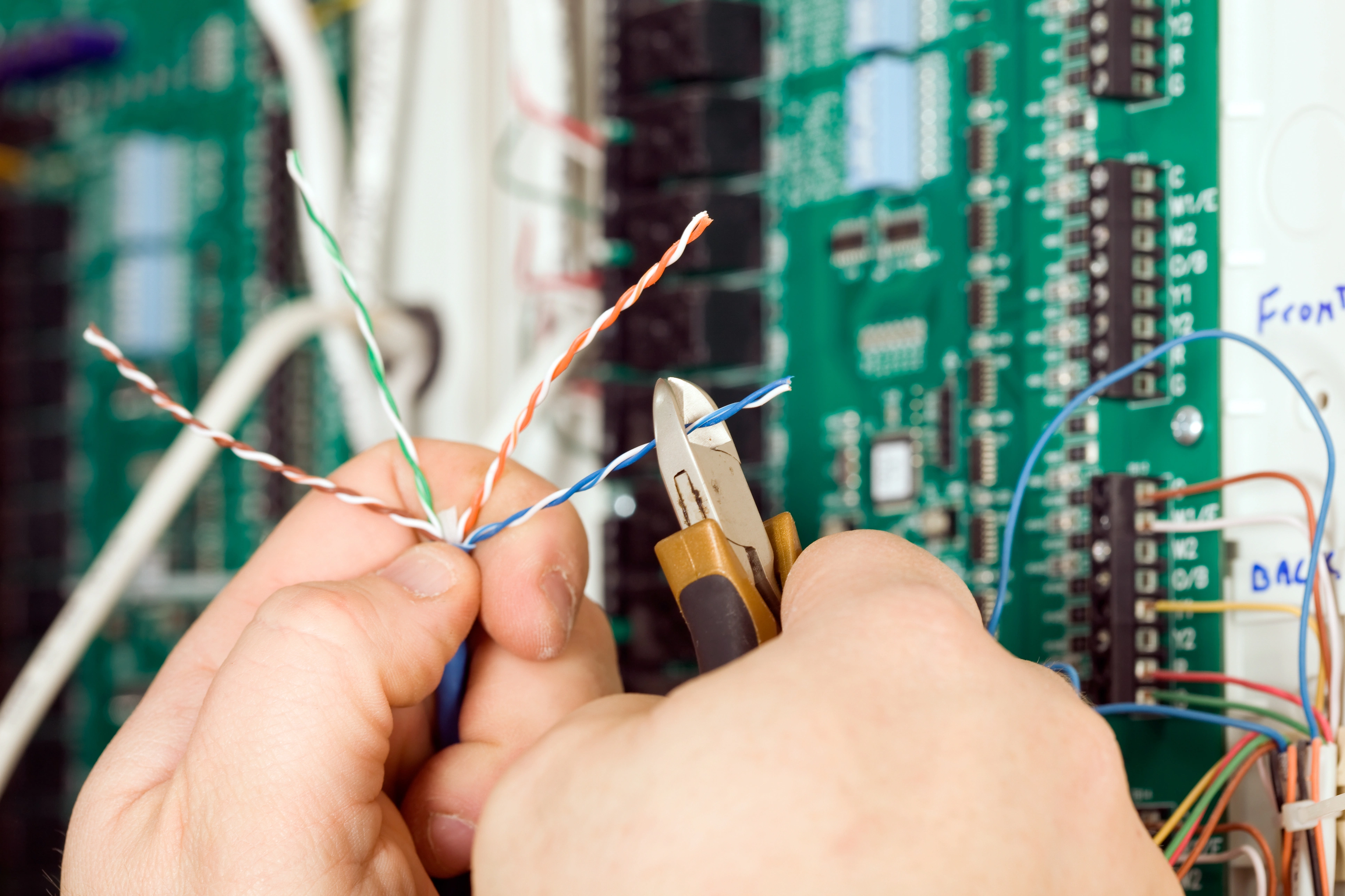 Hands hold pliers, cutting twisted electrical wires against a backdrop of circuit boards.