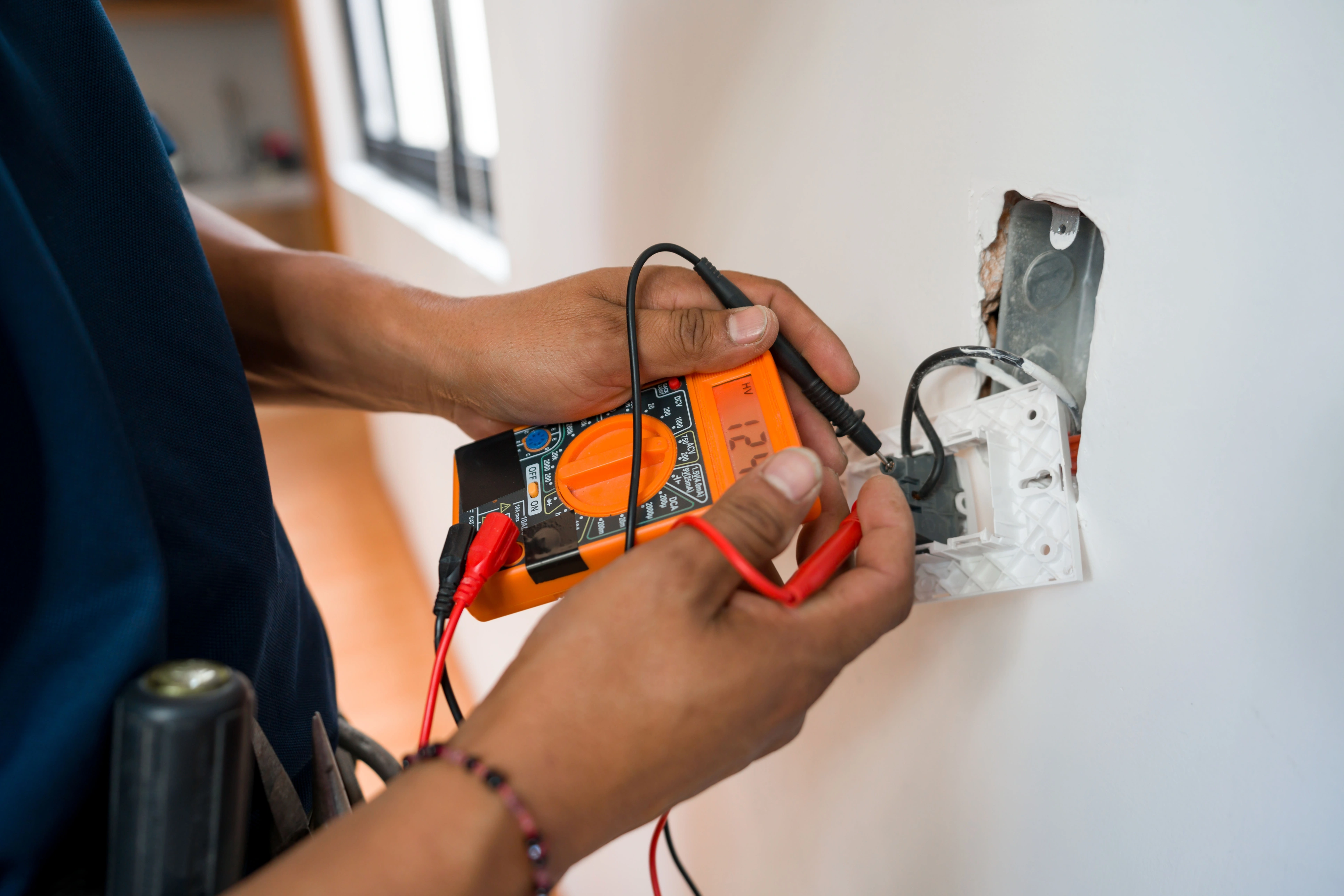 A person uses a digital multimeter to test electrical wires in a wall socket.