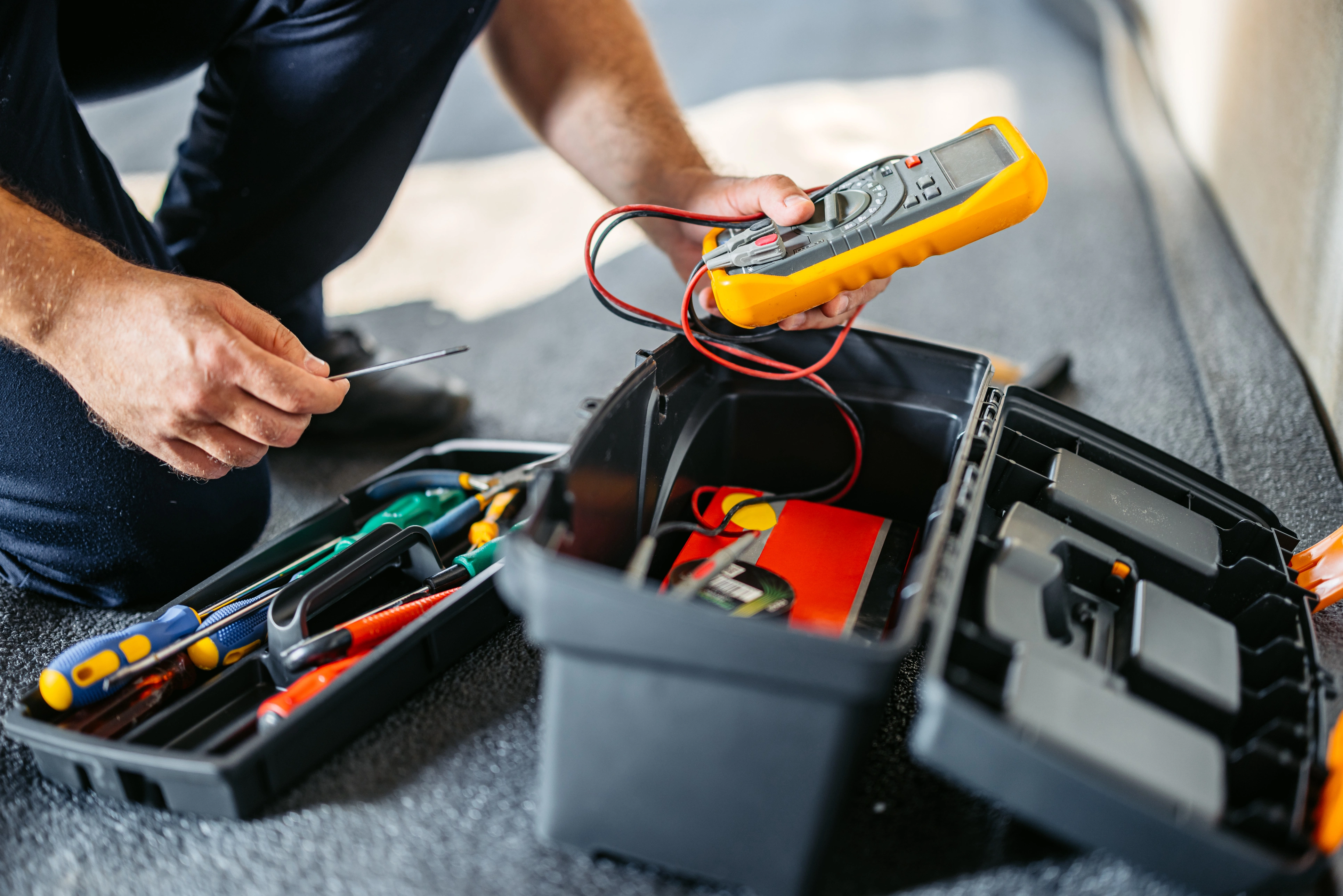 A person uses a multimeter from a tool kit, featuring various tools like screwdrivers and wrenches on a textured surface.