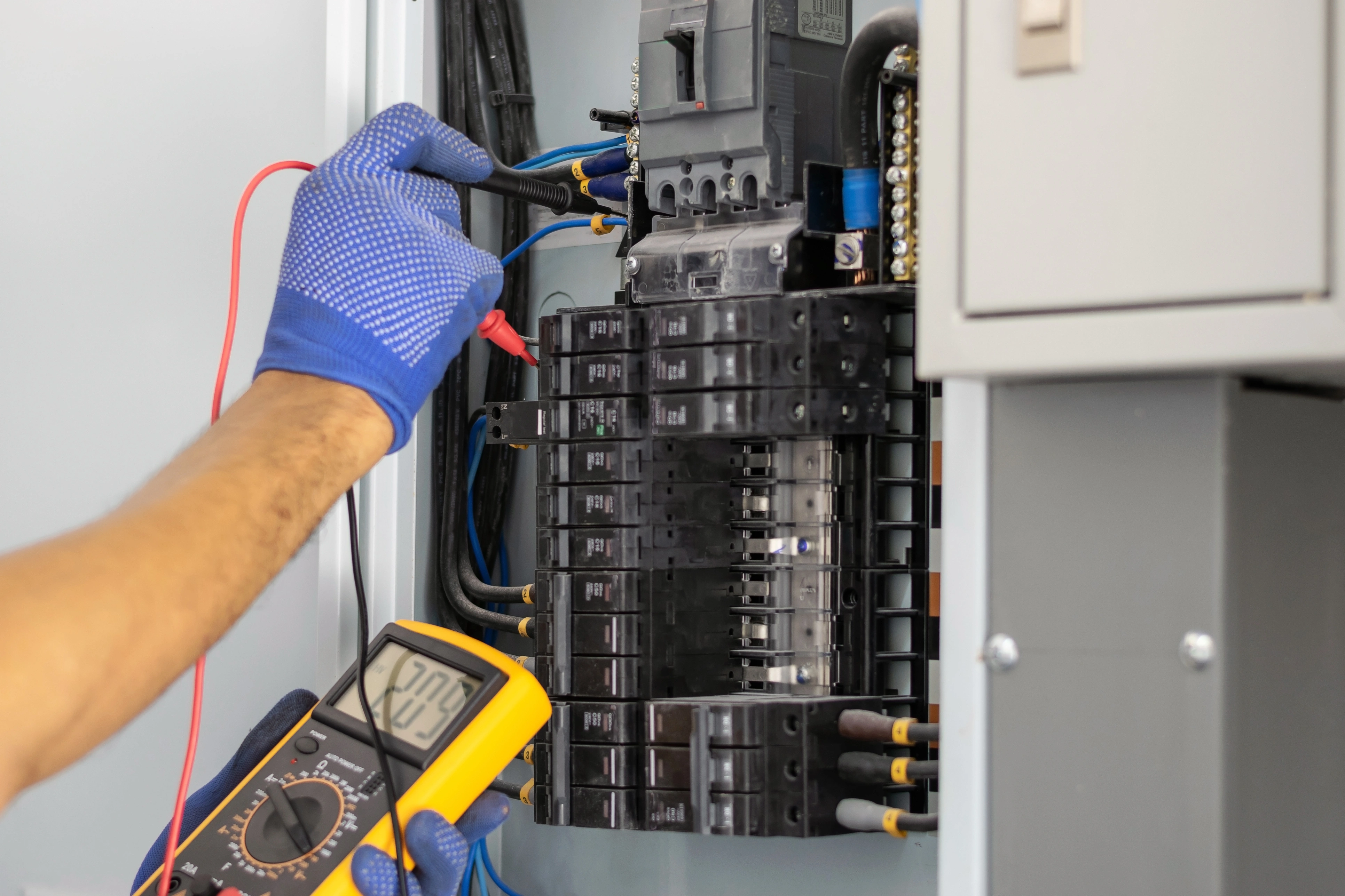 Electrician in blue gloves uses a multimeter to test voltage in a circuit breaker panel.