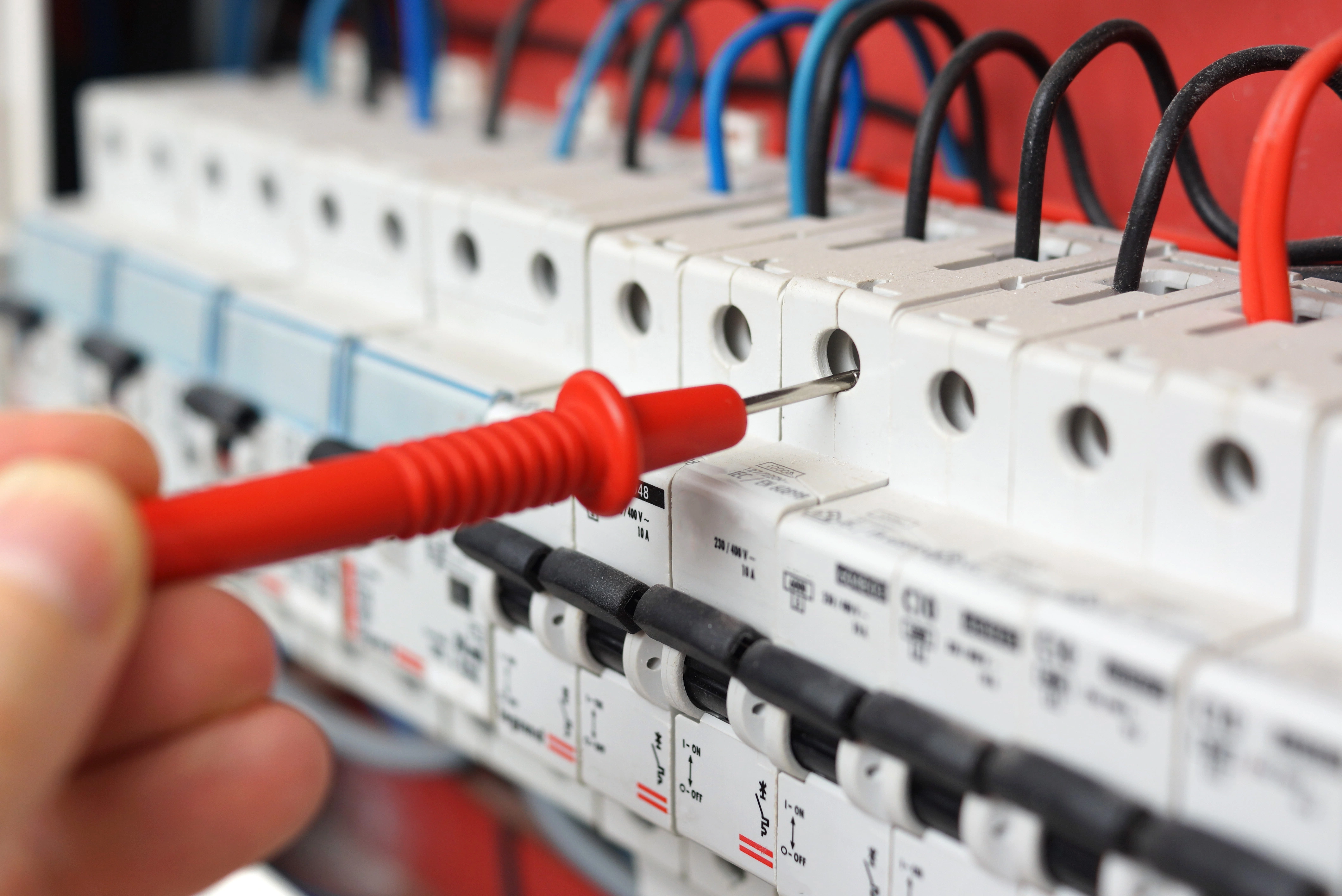 Close-up of a person using a red multimeter probe to test a row of white electrical circuit breakers.