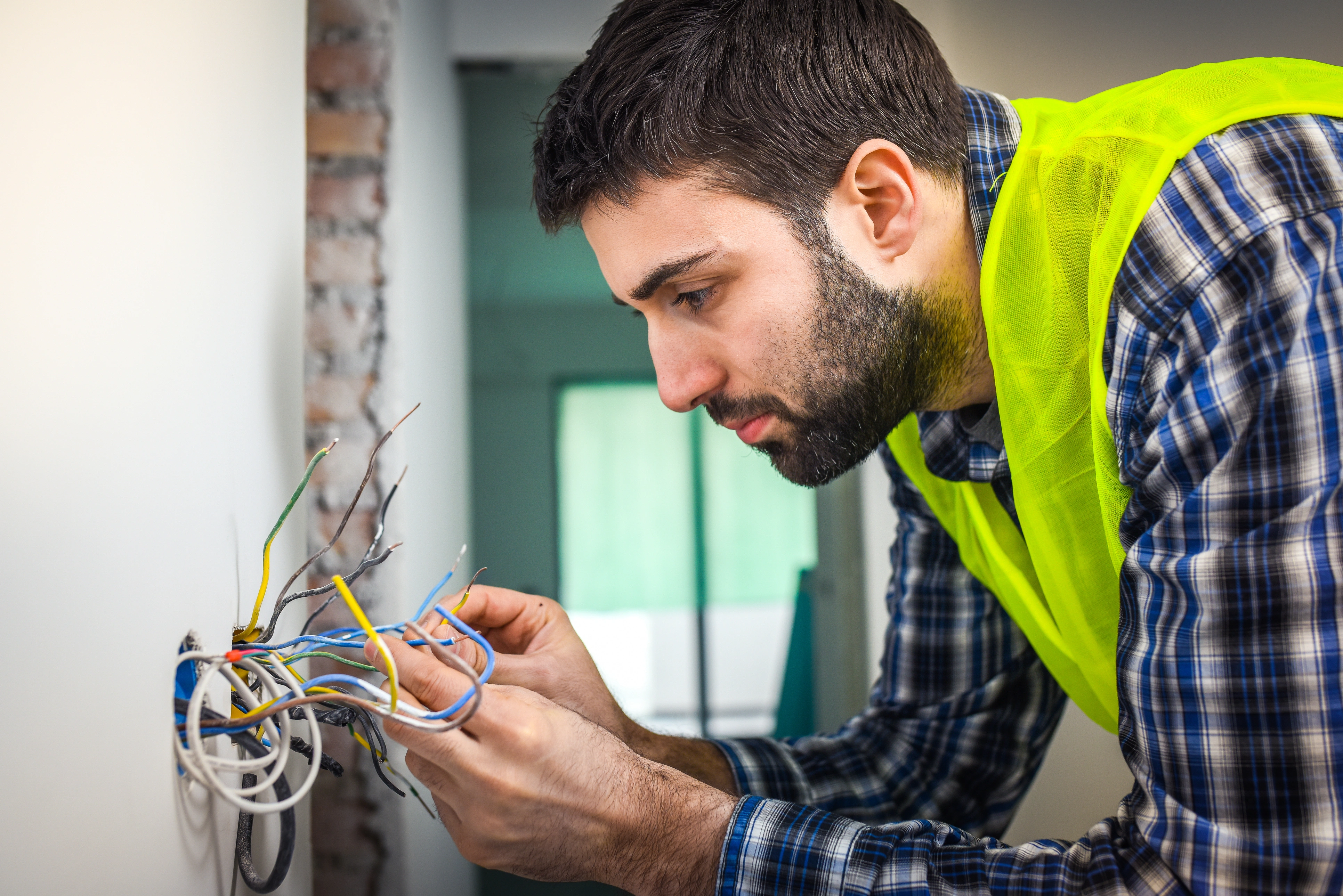A focused electrician in a plaid shirt and yellow vest examines multiple exposed wires protruding from a wall.
