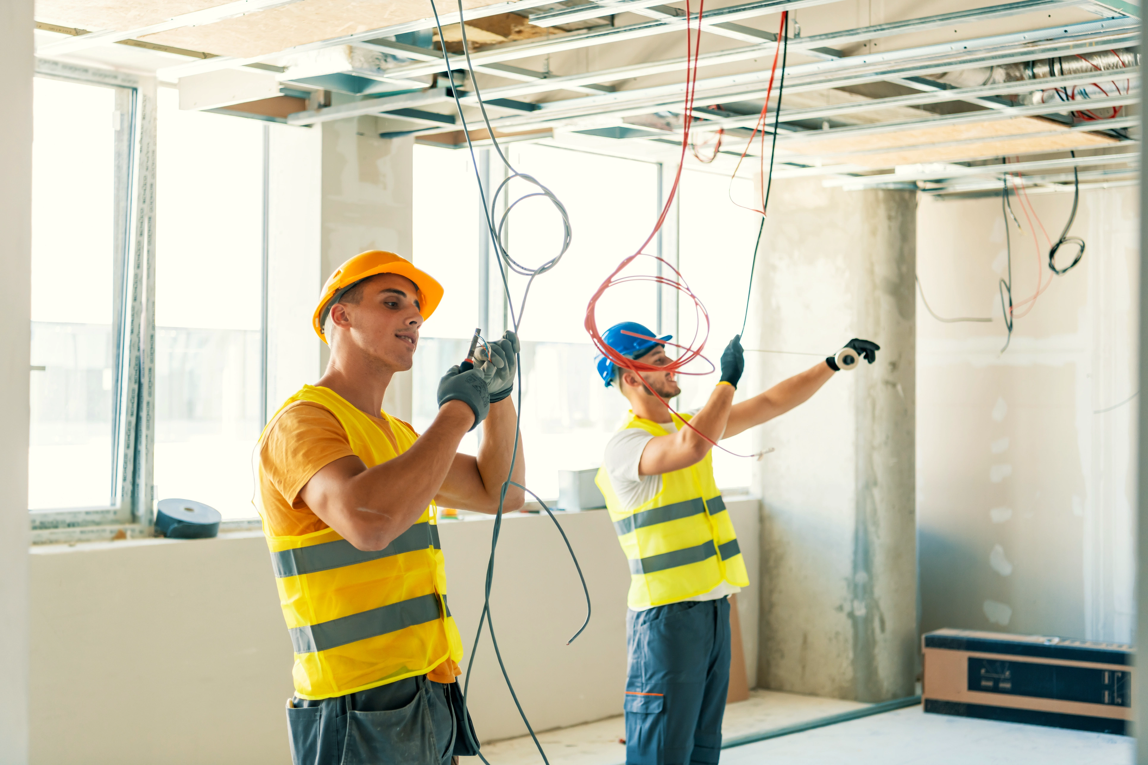 Two construction workers in yellow vests and hard hats work on ceiling wiring in a bright room.