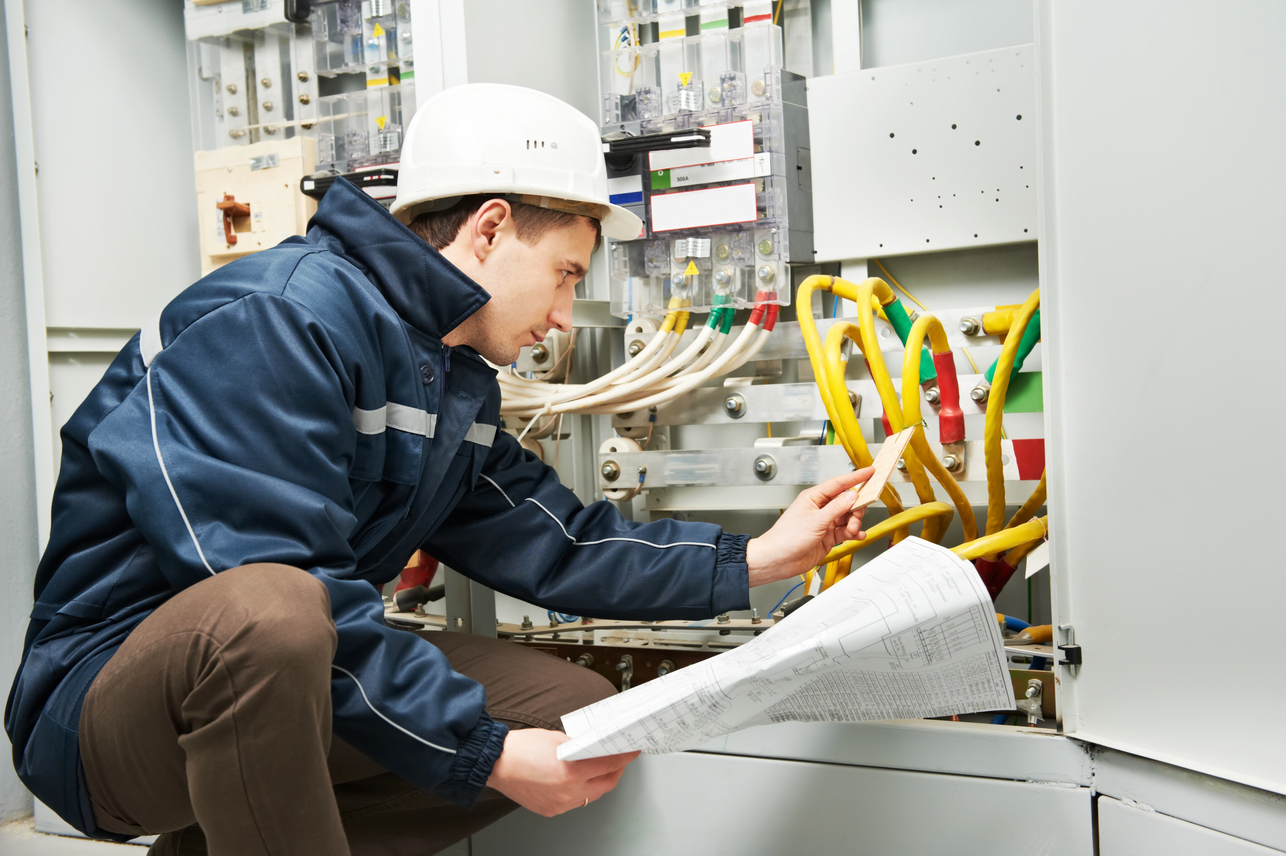 Man in a hard hat and blue jacket examining electrical panels