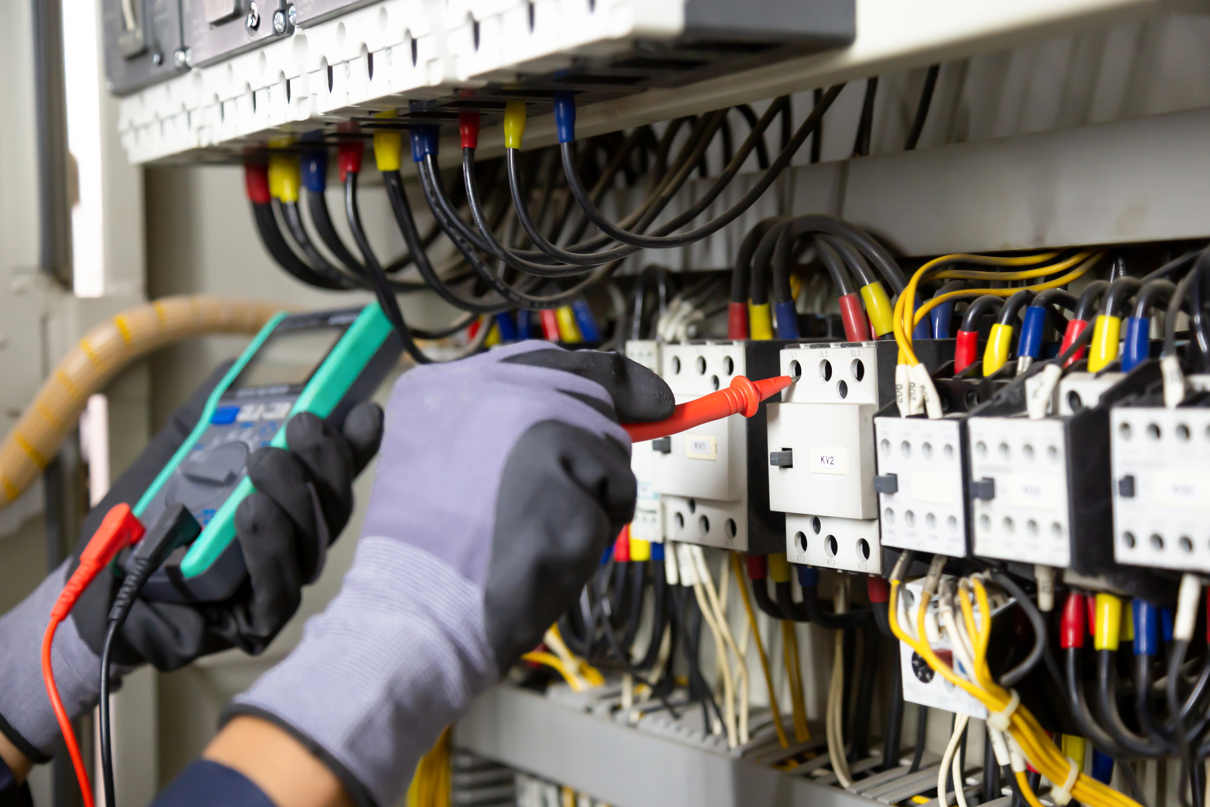 Electrician in gloves uses multimeter to test connections in a circuit breaker panel. 