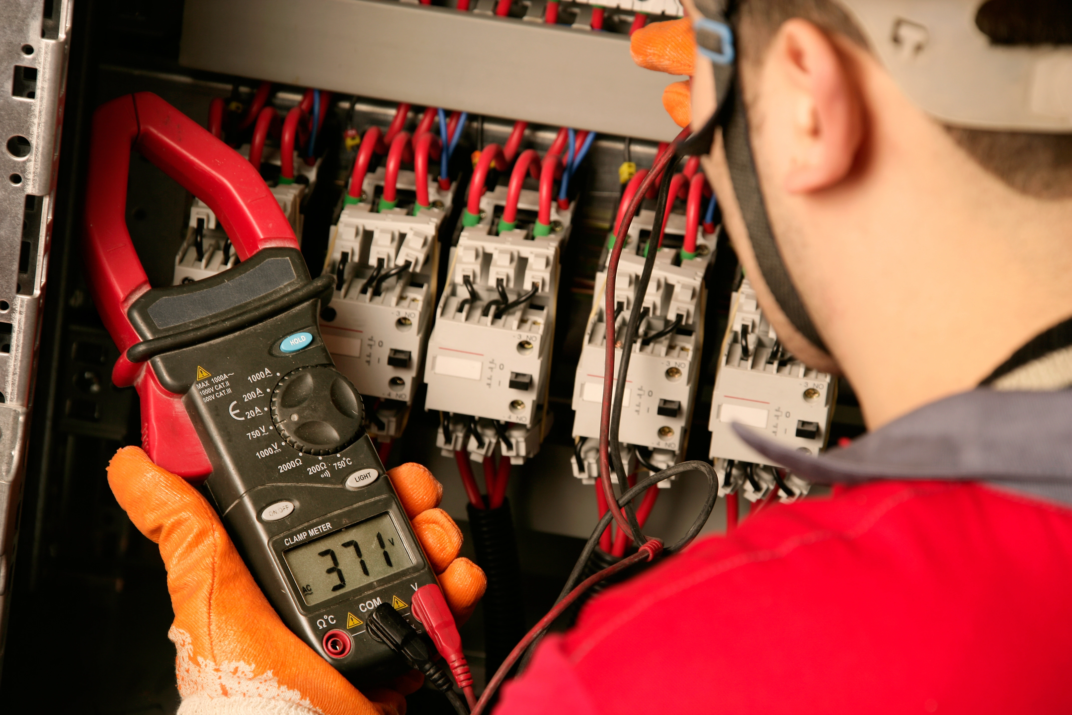 A technician in safety gear uses a clamp meter, to measure electrical current in a panel. 