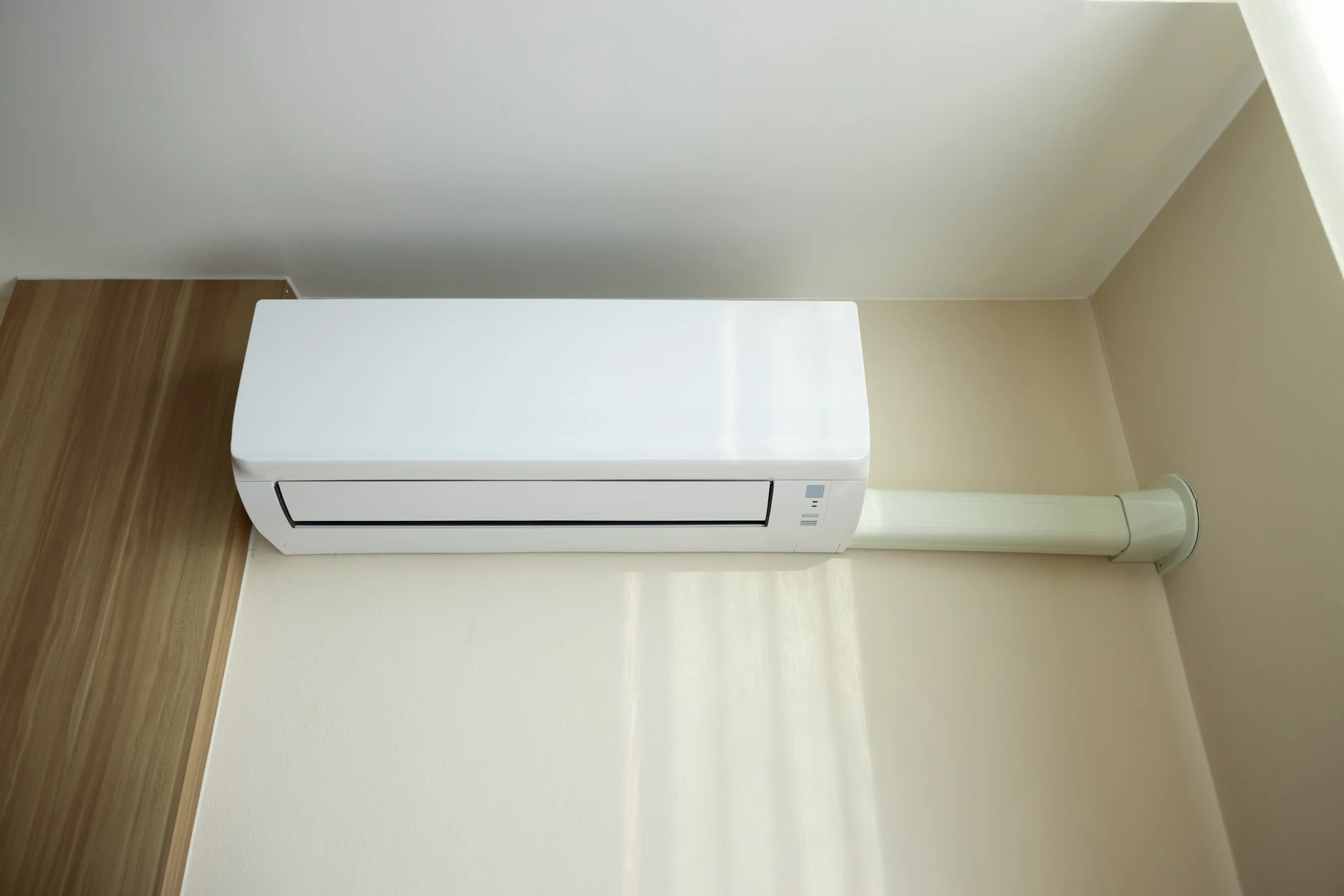 An indoor ductless heat pump is mounted on a beige wall, with a white, insulated conduit running to the right and into the wall.