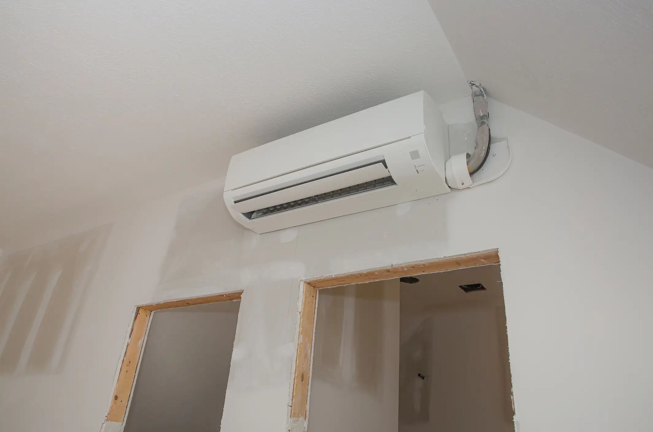  A ductless heat pump, also known as a mini-split, is mounted high on a white wall in a room under construction. The interior unit has a white, rectangular casing. The pipes connecting to the unit are visible, exiting through a hole in the wall. The wall has a rough texture, and there are two unfinished door frames below the unit.