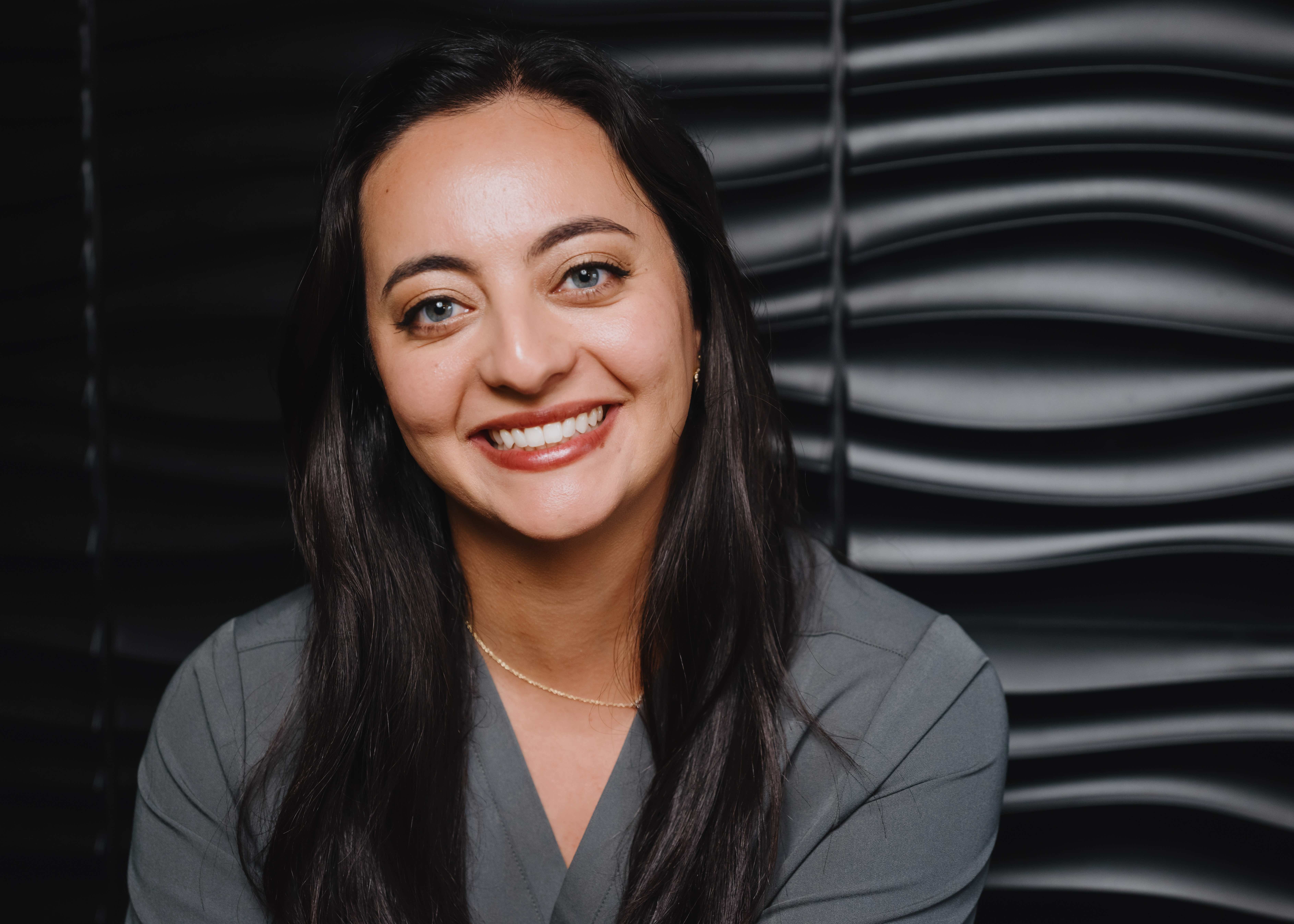 Bald man with a beard, wearing a dark blazer and black shirt, sits on a black leather chair, smiling with hands clasped—radiating the calm confidence of an experienced dentist.