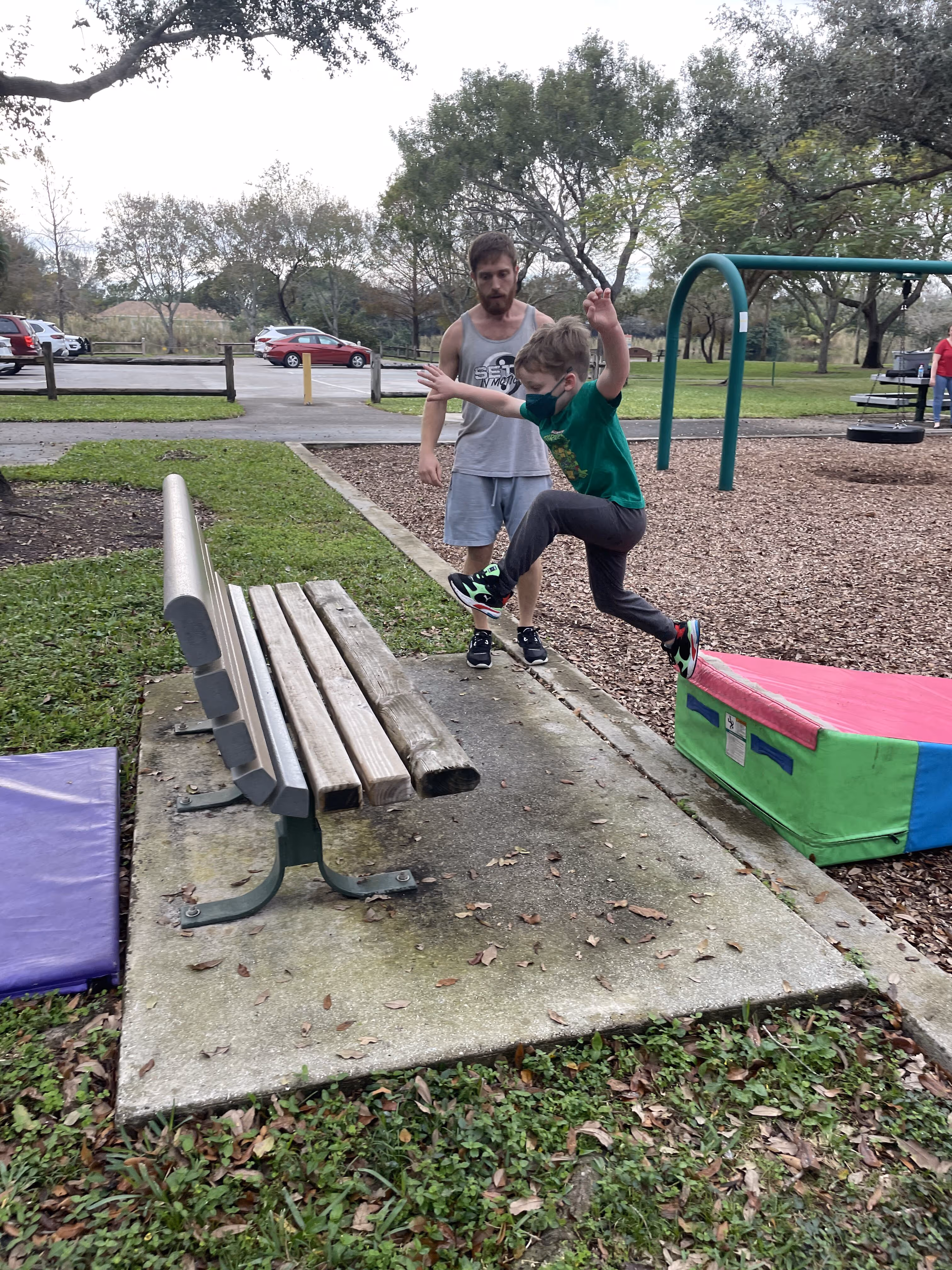 Child wearing a green shirt and face mask jumping off a colorful foam block at a park while a man in a gray tank top watches.
