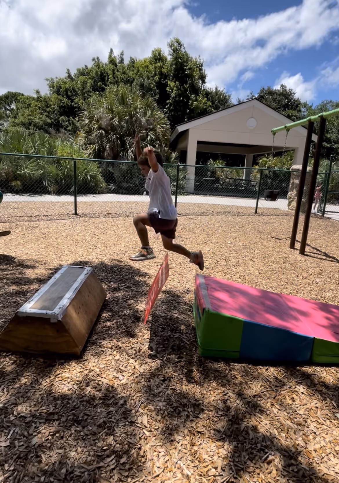 A child in a white shirt and purple shorts jumping over a red sign between a wooden ramp and a colorful foam block in a playground with wood chip ground cover.