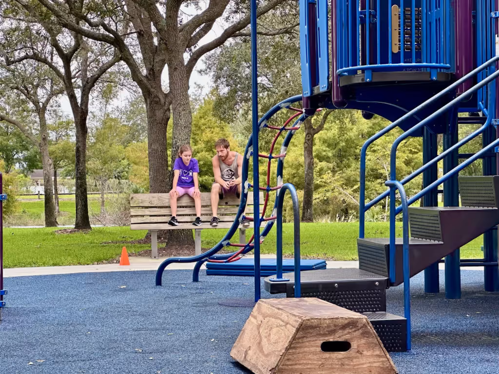 A man and a girl sitting on a park bench near a playground with blue rubber flooring and climbing structures, surrounded by trees and greenery.