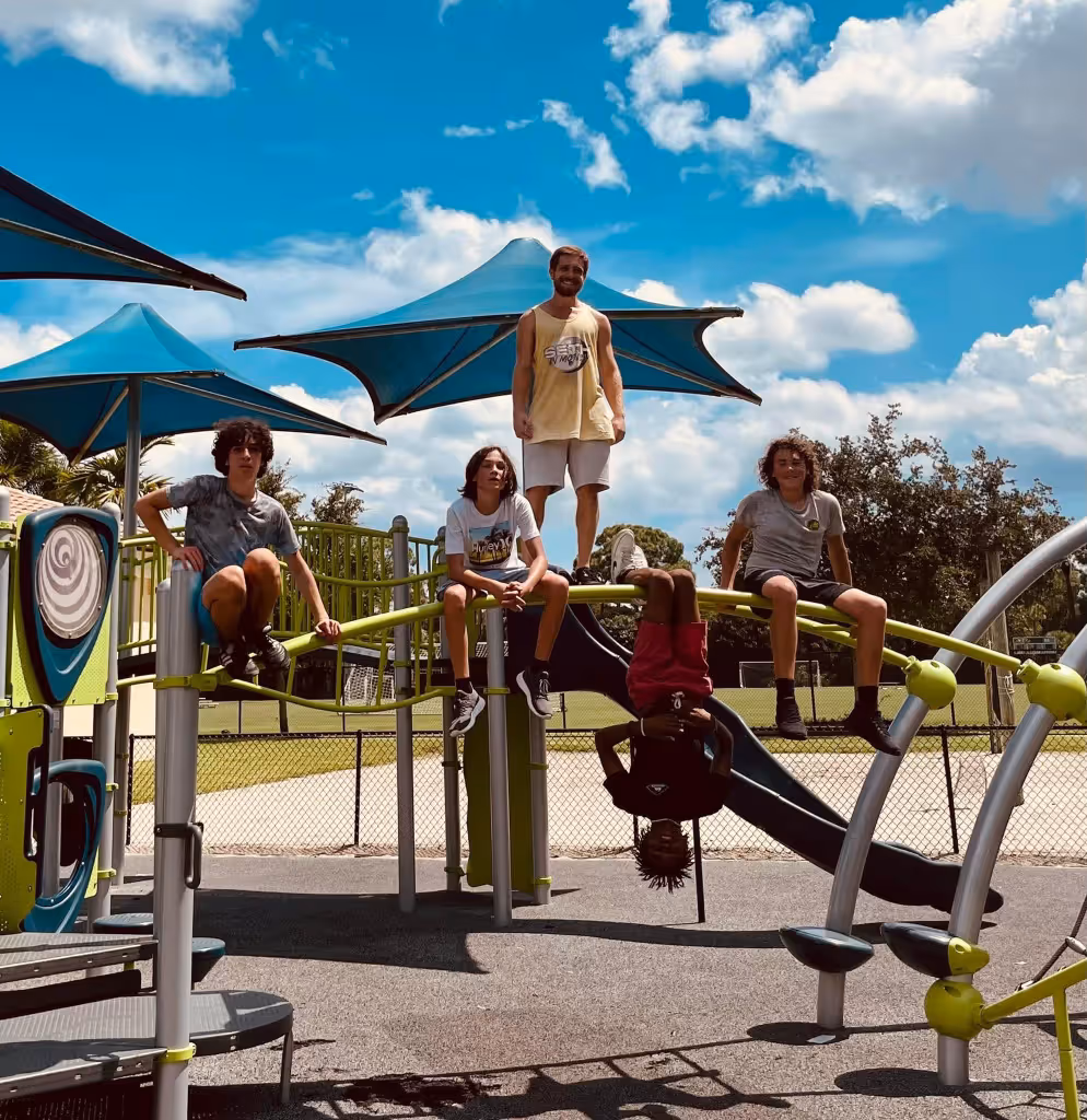 Five young people on playground equipment under blue sky with clouds, one hanging upside down.