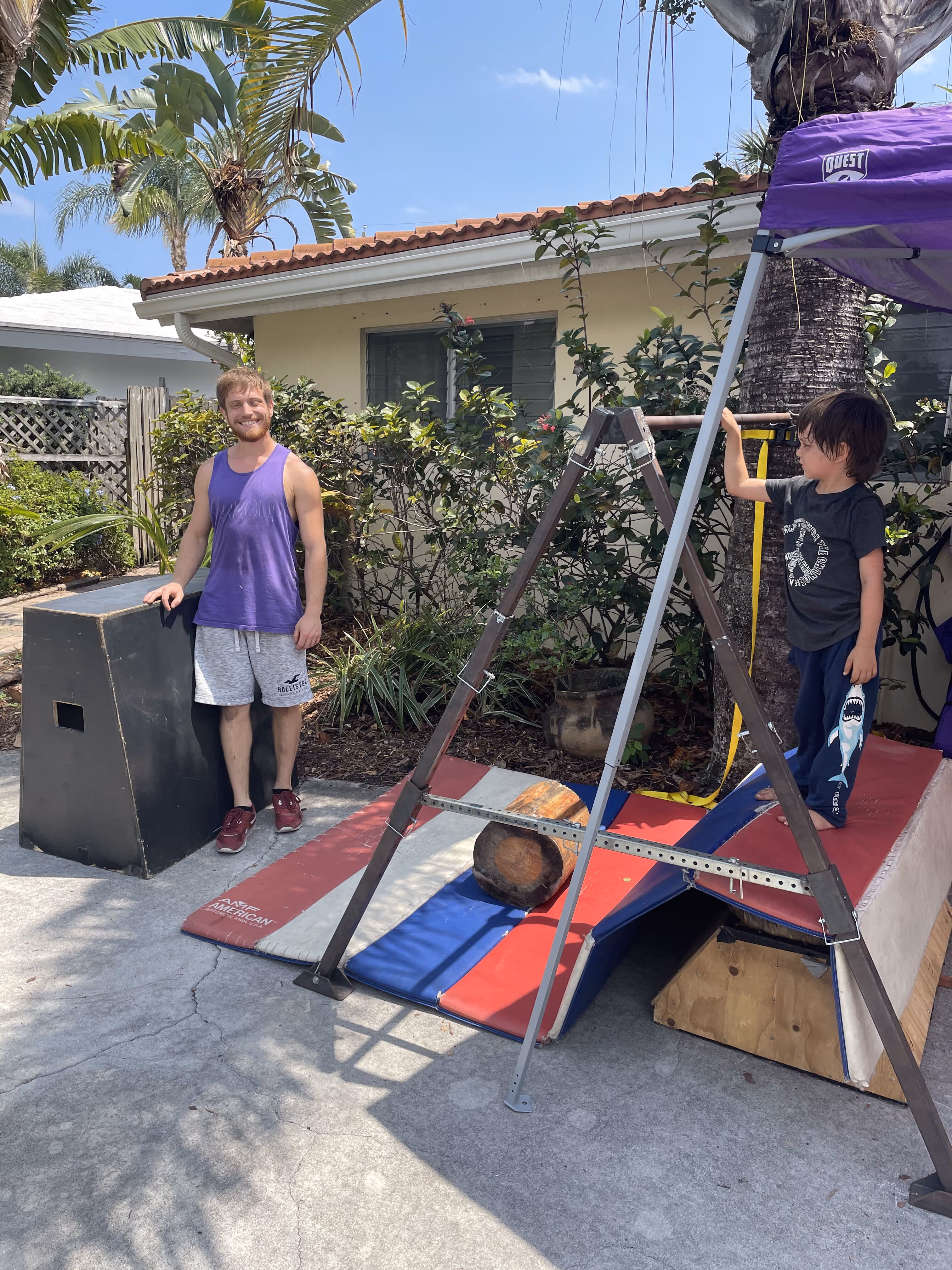 Man in purple tank top standing next to a black box and a child holding a bar on a small outdoor gym setup with mats and a wooden log.