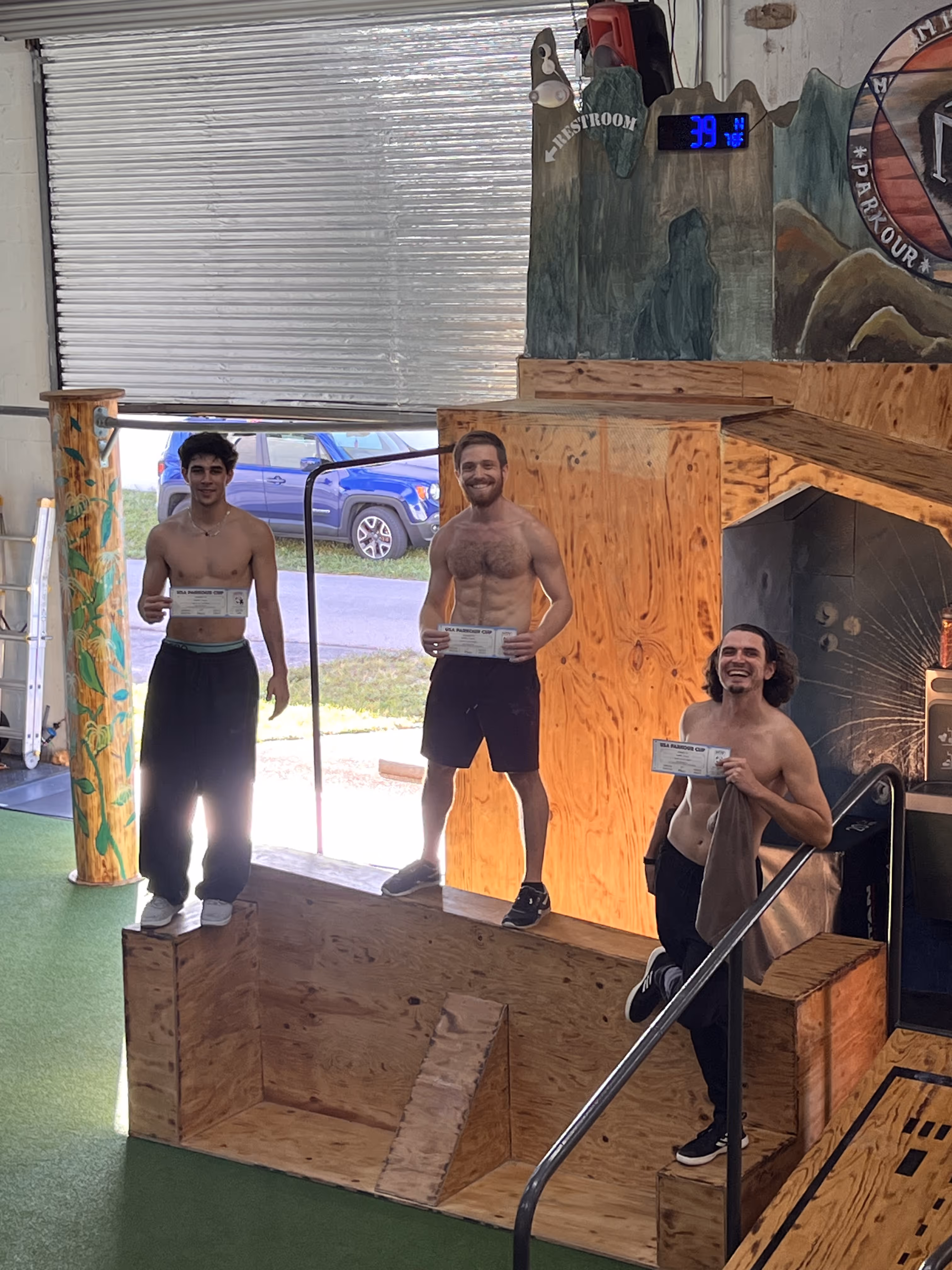 Three shirtless men smiling and holding certificates while standing on a wooden podium inside a gym with parkour-themed walls.
