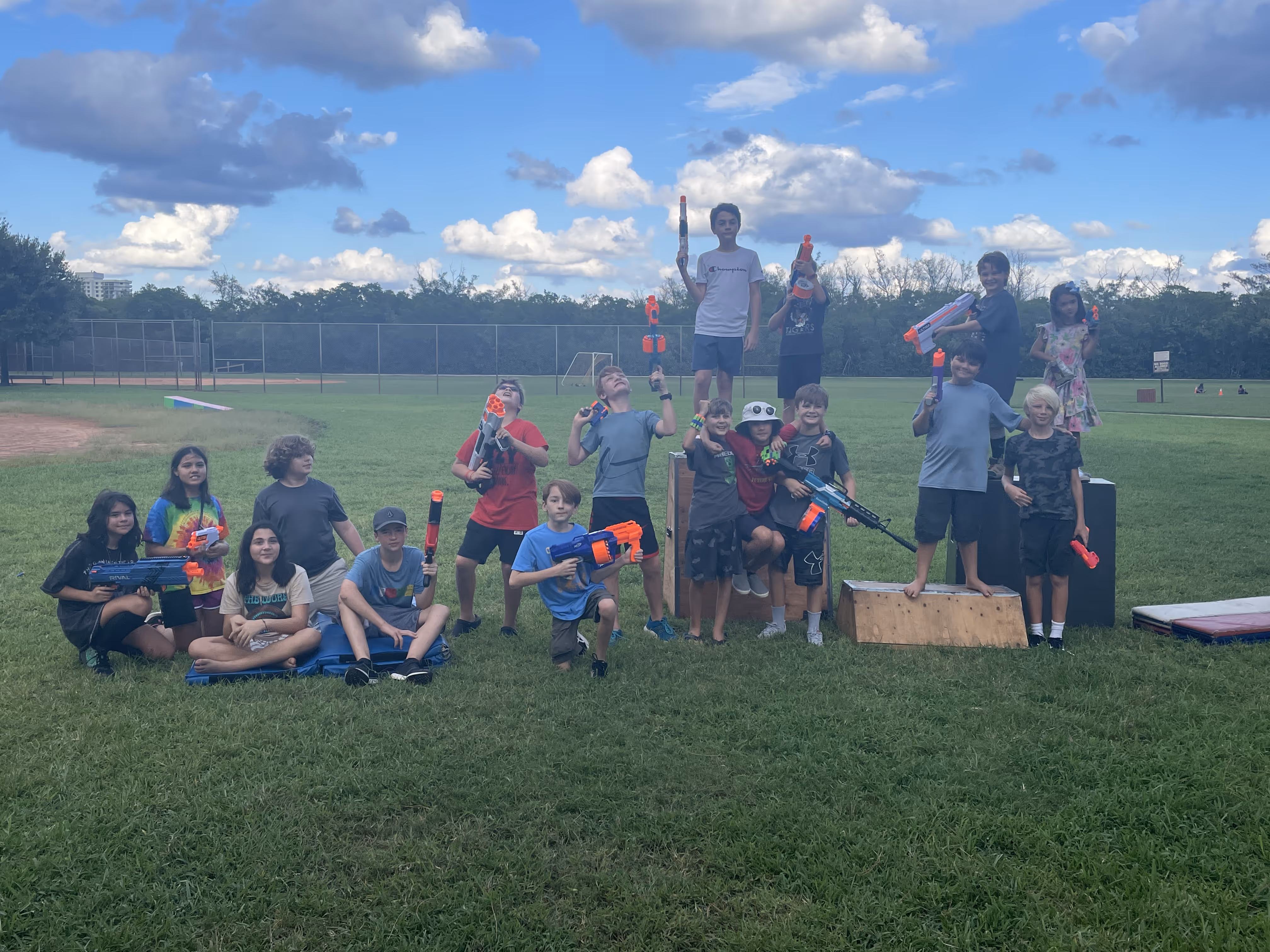 Group of children outdoors on grass holding toy Nerf guns, posing and smiling under a partly cloudy sky.