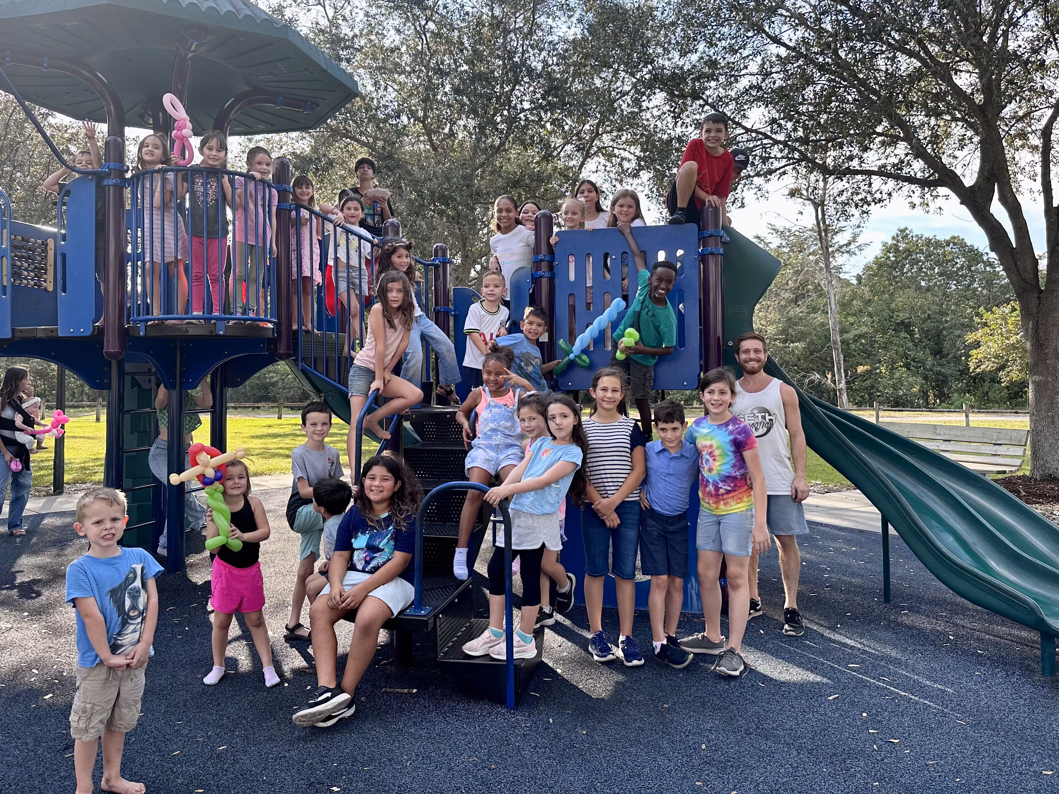 A diverse group of children and one adult smiling and posing on a playground structure with a slide in a sunny park.