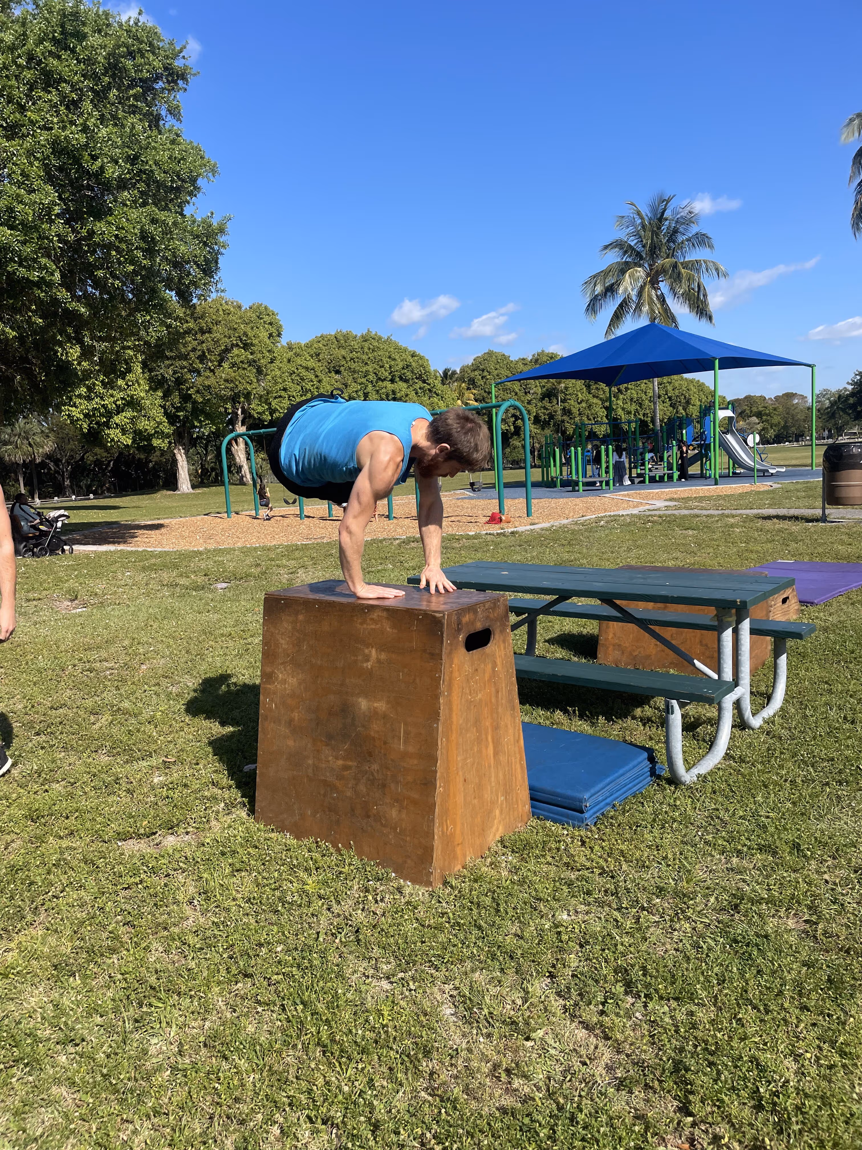 Man wearing a blue tank top balancing on his hands on a wooden box in a park with playground equipment and trees in the background.