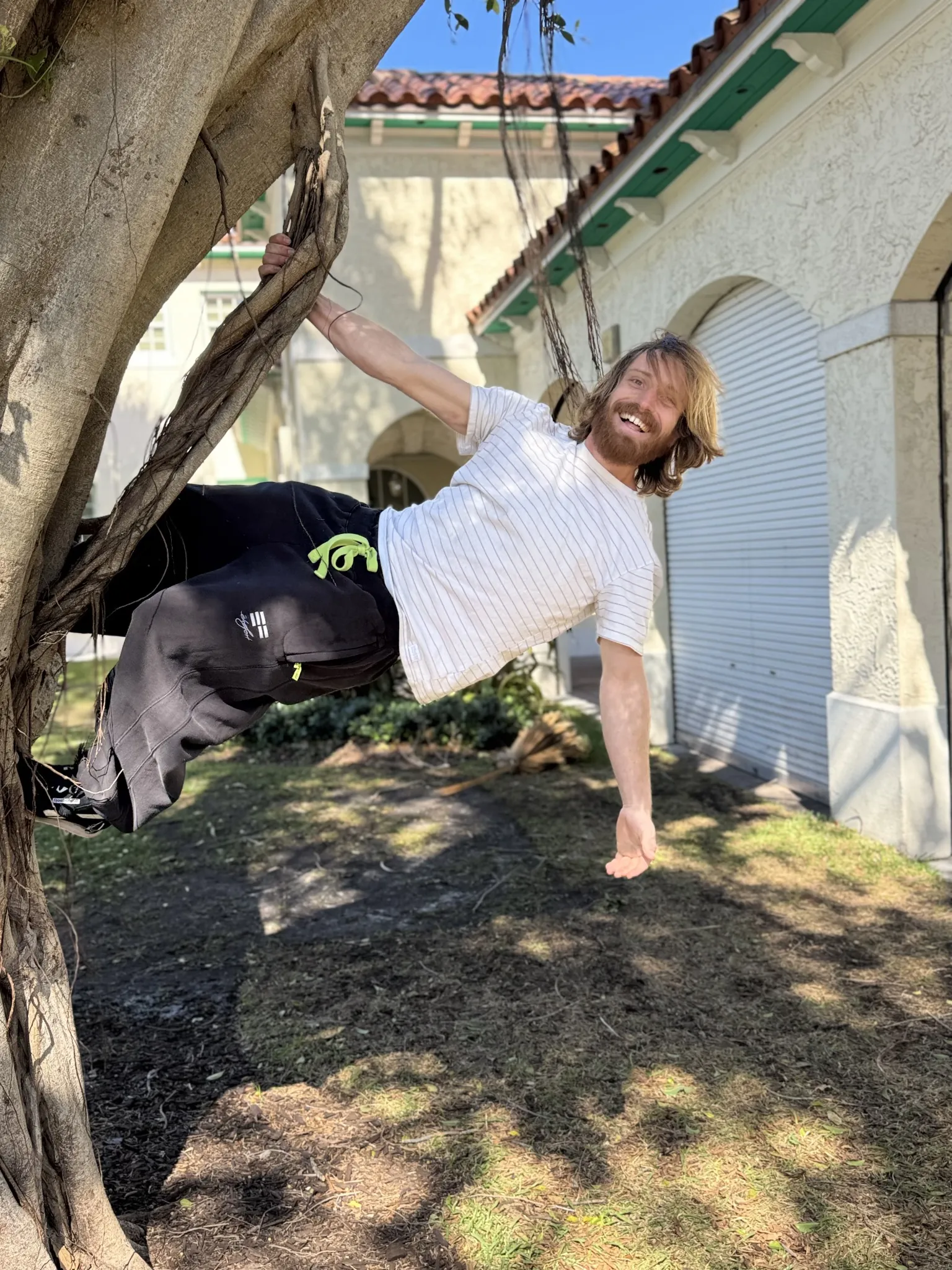 Seth with long hair and beard wearing a white striped t-shirt and black pants hanging sideways from a tree branch, smiling outdoors near a building with arched doorways.