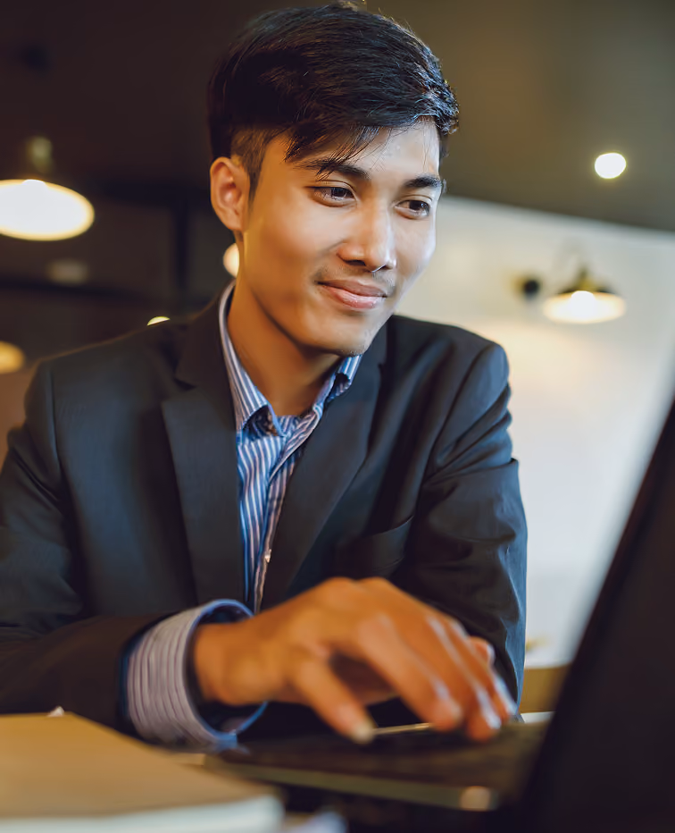 Young man in a suit jacket and striped shirt sitting indoors, smiling and working on a laptop.