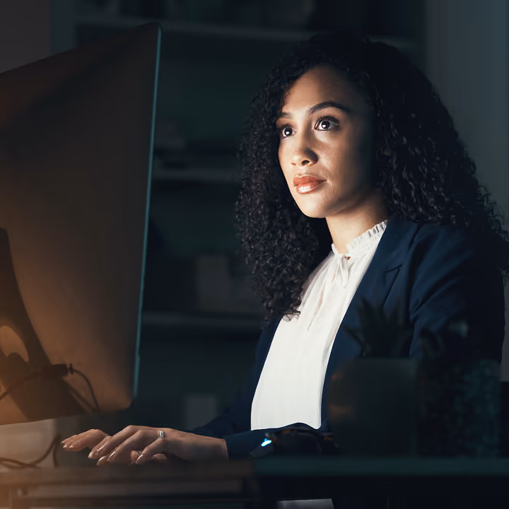 A woman with curly hair wearing a white blouse and dark blazer working intently on a computer in a dimly lit room.