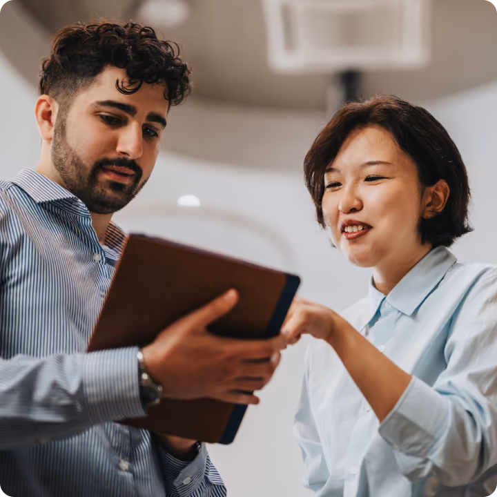 A man with curly hair and a beard wearing a striped shirt looks at a tablet while a woman with short hair in a light-colored shirt points at the screen in an office setting.