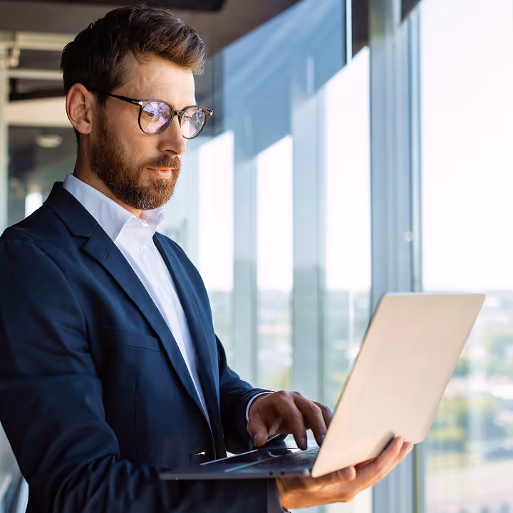 Bearded man in glasses and a dark suit holding and working on a laptop near large office windows with a city view outside.