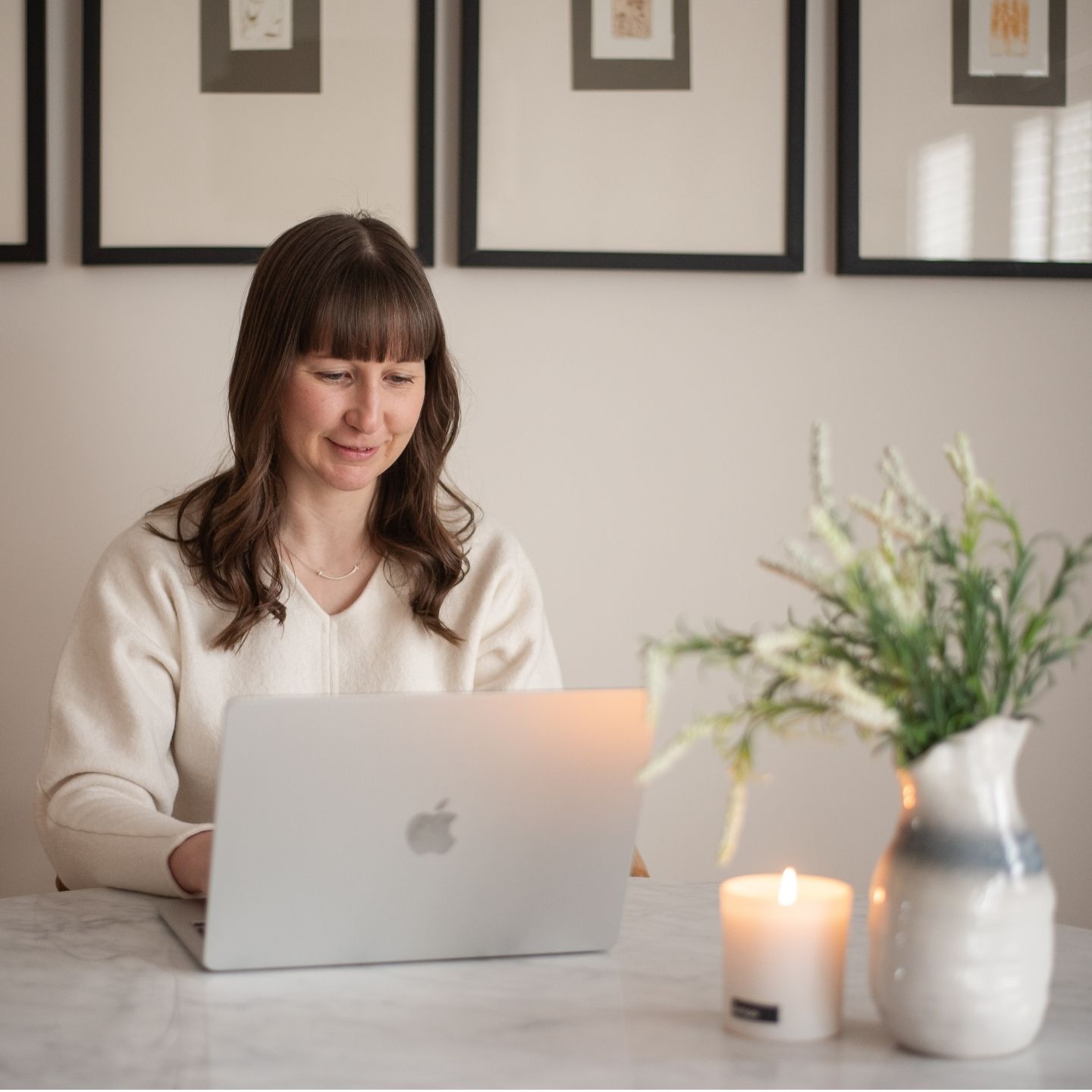 Megan Cook looks at her laptop while smiling. She is sitting at a marble table that has a tempo candle and plant on it. 
