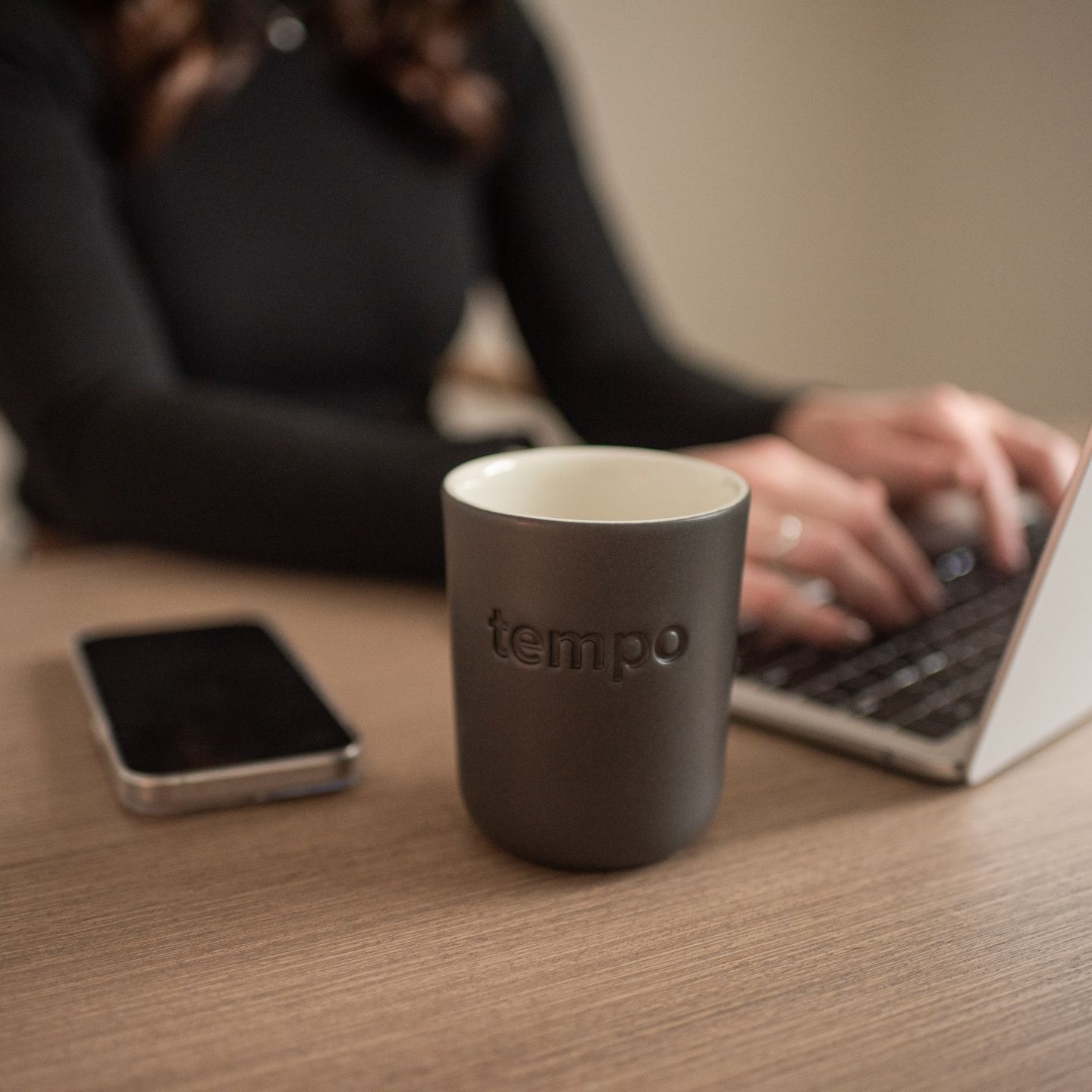 A tempo mug and phone are on a desk with Megan Cook typing on a laptop in the background..