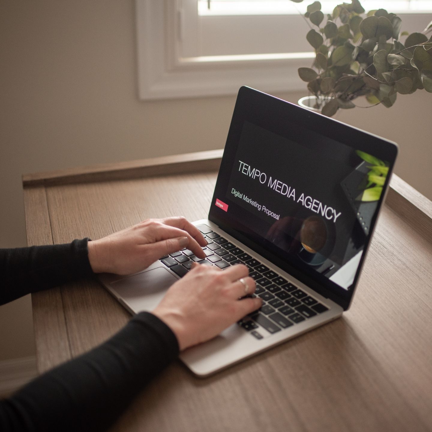 hands typing on a laptop that is on a brown desk. the screen shows a tempo media agency digital marketing proposal on the screen. 