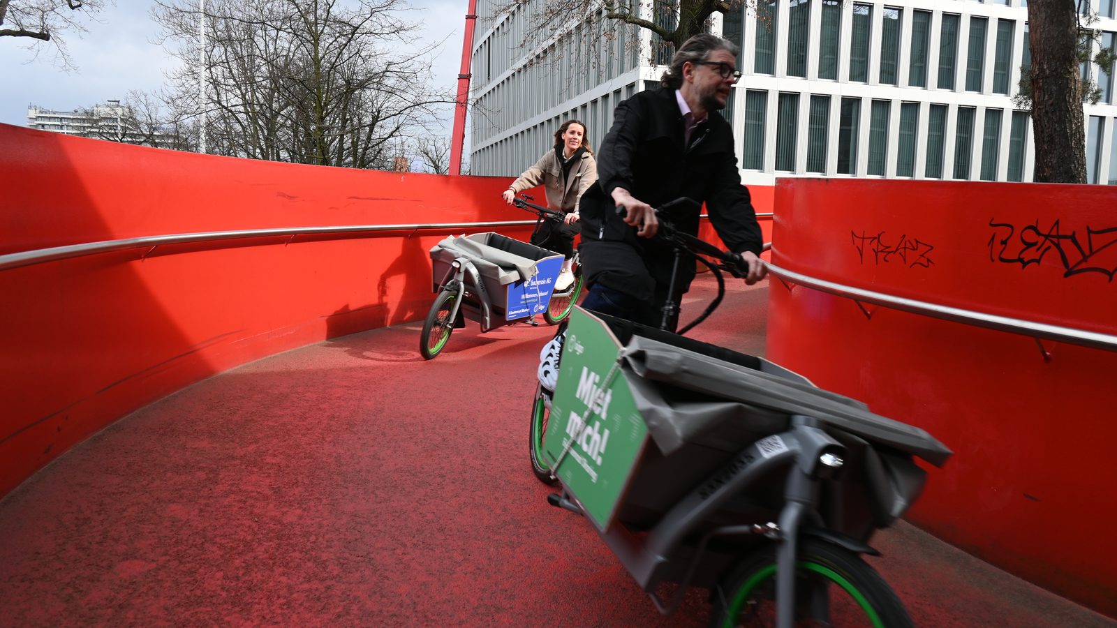Two riders on sigo e-cargo bikes crossing a bright red cycle bridge in a city
